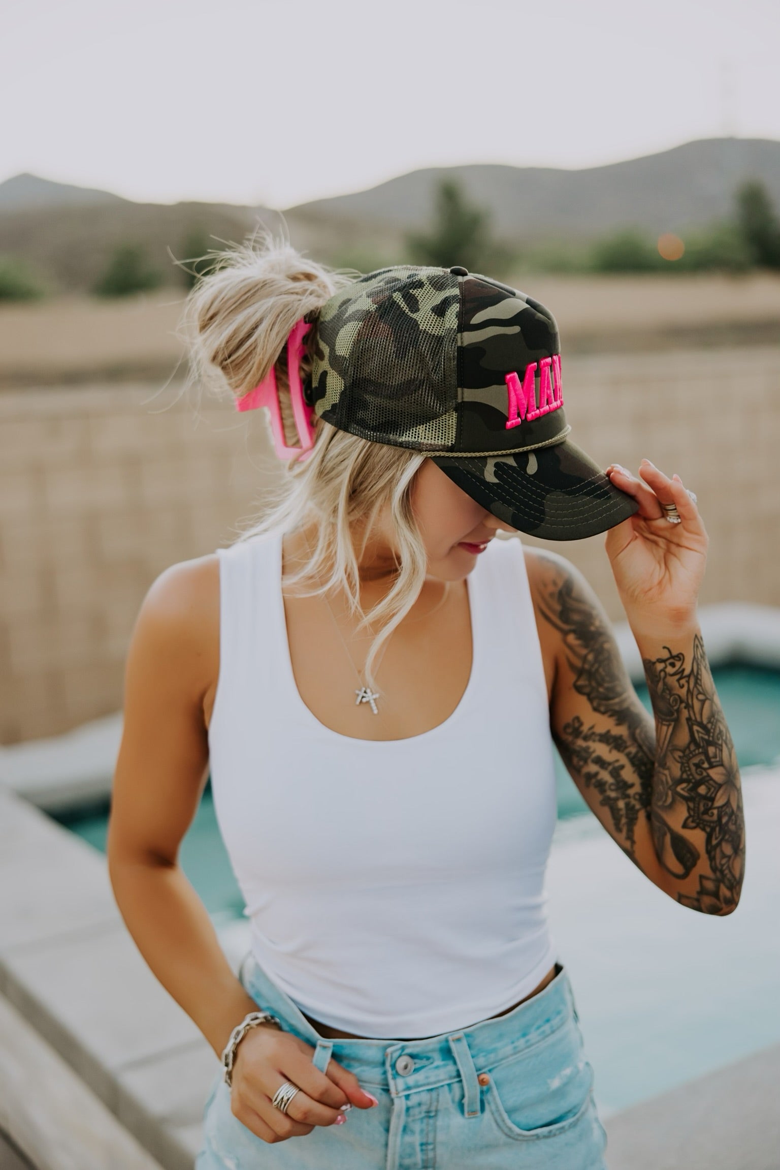 A woman in a white tank top and denim shorts, with a tattooed arm, adjusts her camouflage cap with "MAMA" in pink letters, standing by a pool with mountains in the background.