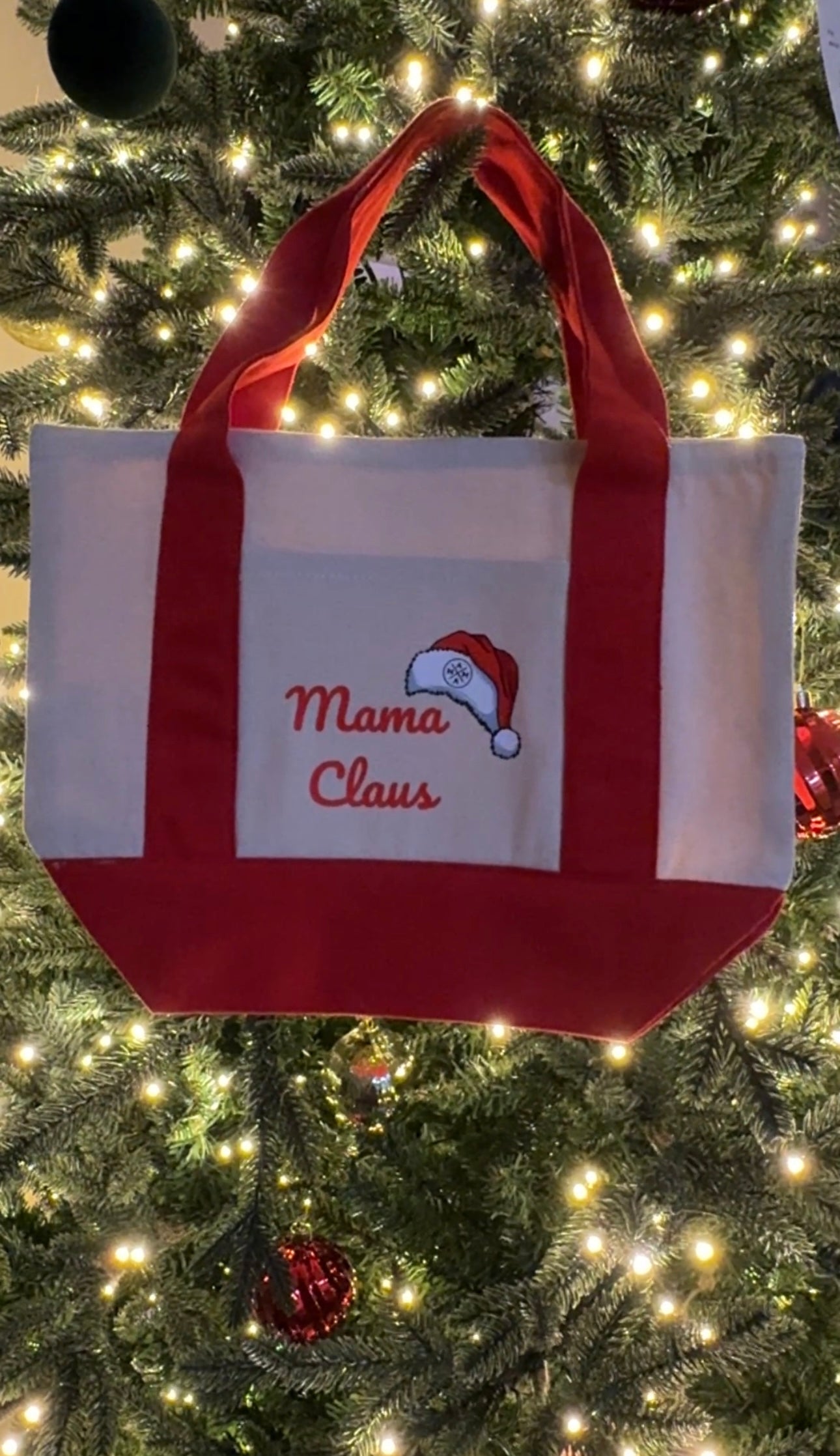 A cream and red tote bag with “Mama Claus” and a Santa hat embroidered on it hangs in front of a decorated, lit Christmas tree.