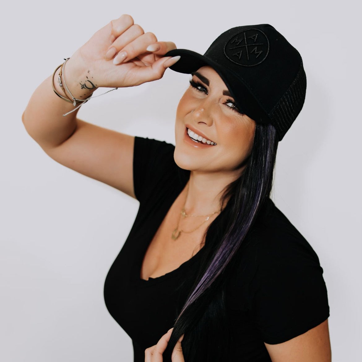 A smiling woman with long dark hair wearing a black T-shirt and black cap holds the cap’s brim with one hand. She has bracelets on her wrist and is posing against a plain white background.