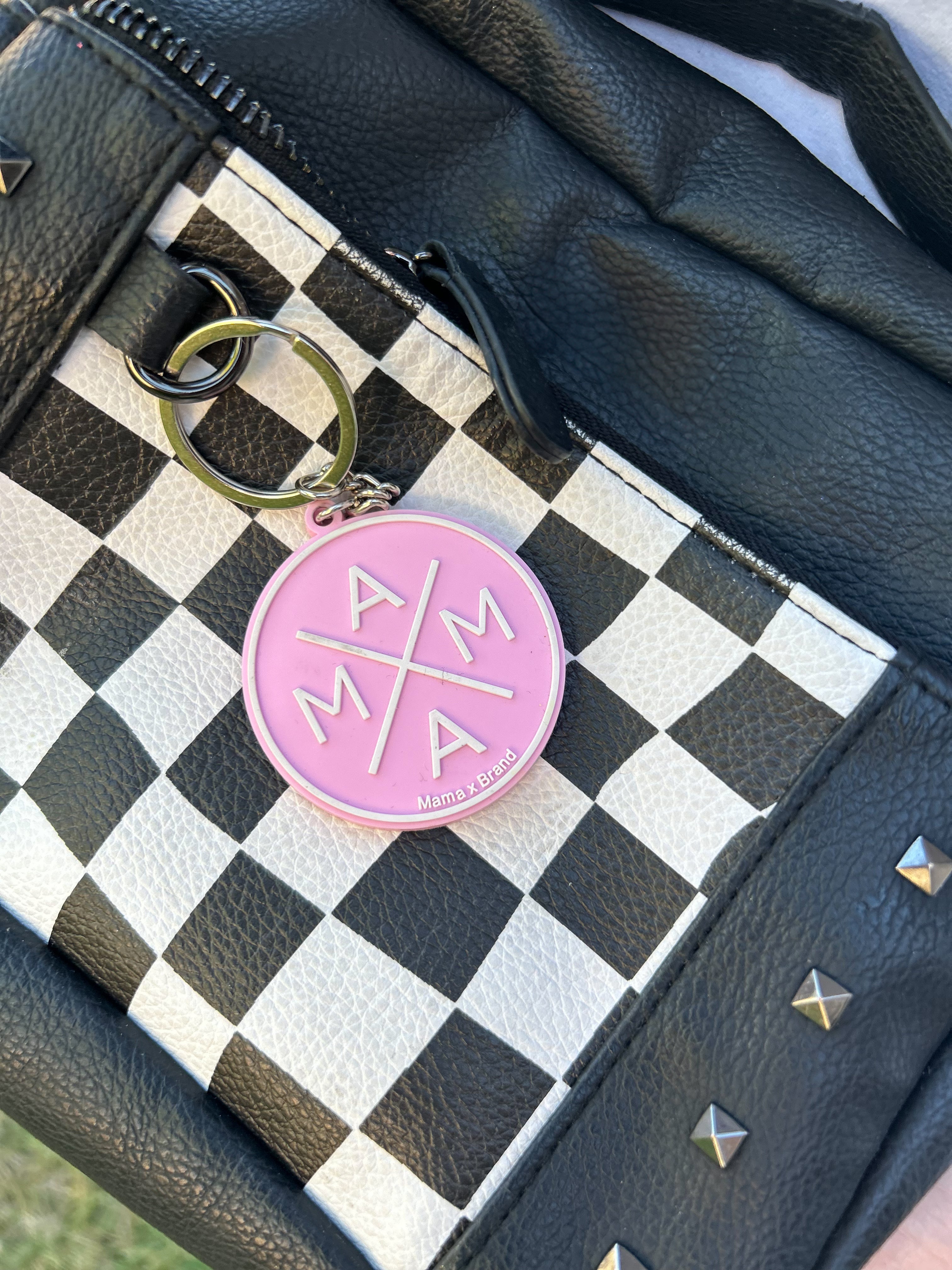 The Mama X™ Keychain by Mama X™ Brand, in pink with a gold ring, sits atop a black-and-white checkered diaper bag with black leather trim and metal studs—an ideal accessory for moms.