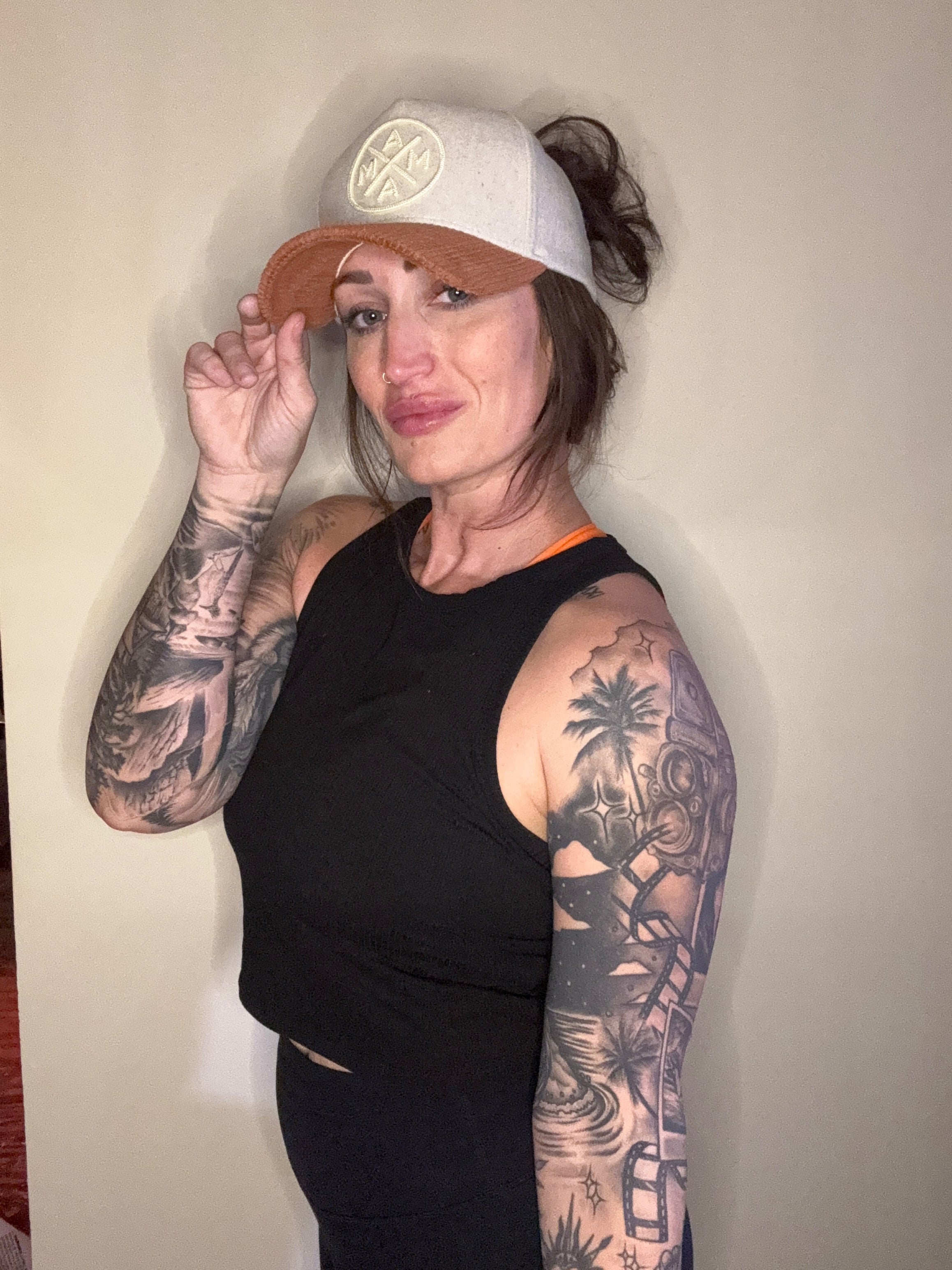 A woman with long brown hair in a bun, wearing a sleeveless black crop top and detailed tattoos on her left arm, smiles while holding the brim of Courtney’s Trucker Hat by Mama X™ Brand against a plain light wall.