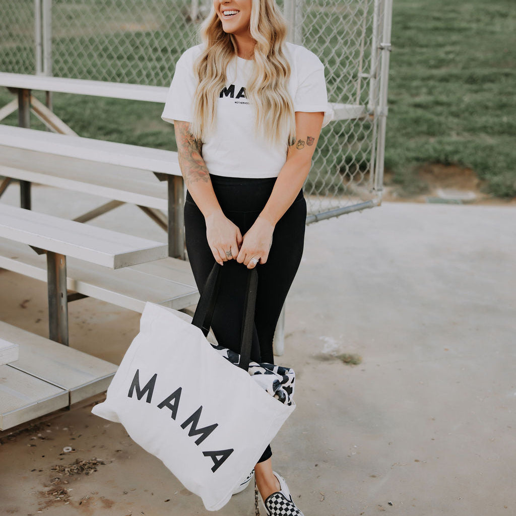 A woman with long blonde hair smiles near bleachers, wearing a white "MAMA" t-shirt, black pants, checkered shoes, and carrying the oversized Mama Tote from Mama X™ Brand—an ideal bag for moms. Grass and a chain-link fence are in the background.