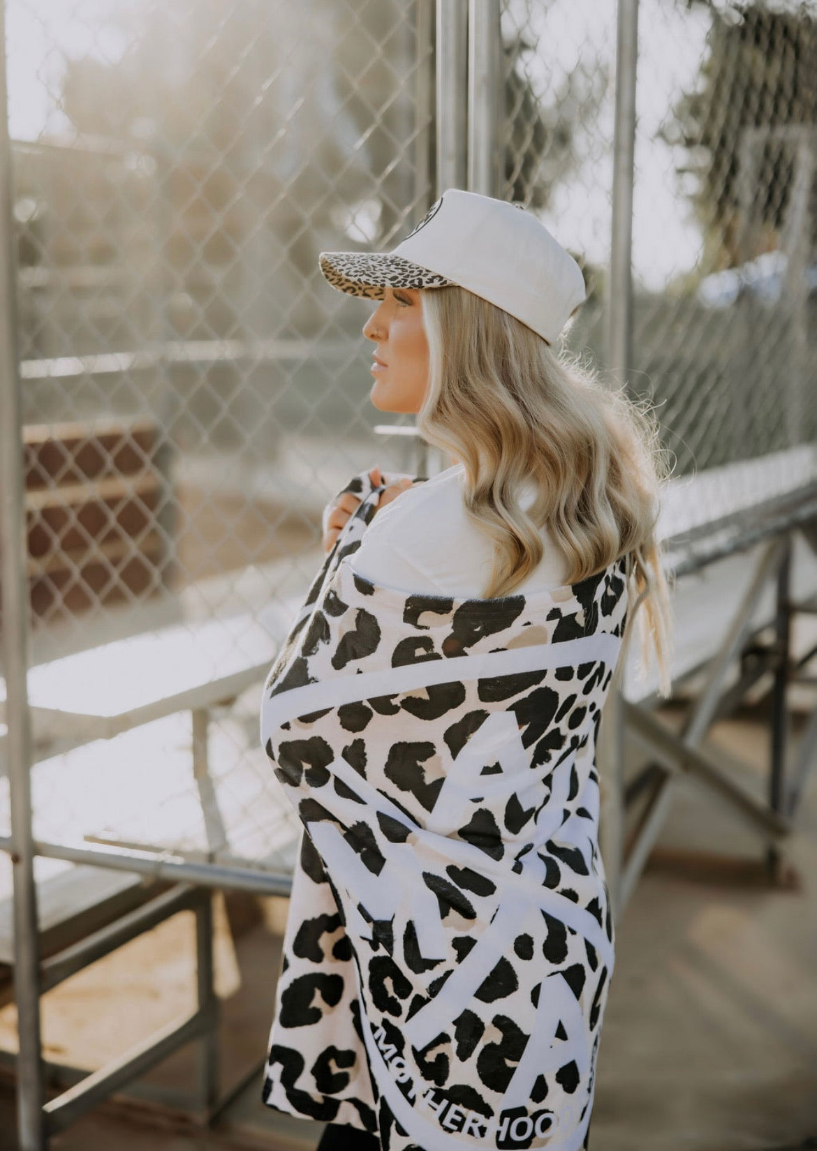 A woman with long blonde hair, sporting a white cap and wrapped in a Mama X™ Brand Motherhood Is So Fetch Towel, stands by metal bleachers near a chain-link fence on a sunny day.