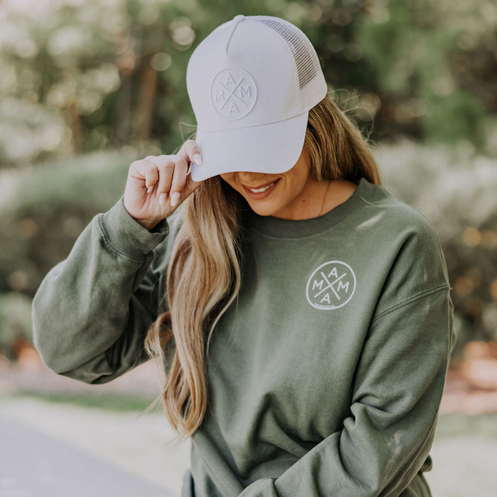 Sitting outside on a stone surface surrounded by greenery, a person in a green sweatshirt and black shorts adjusts their Mama X™ Whiteout Premium Trucker Hat, showcasing the iconic logo. They have long hair flowing freely as they enjoy the serene outdoors.