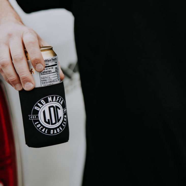 A hand is holding a canned drink wrapped in a black koozie. The koozie, perfect as a gift for Dad, has a white circular logo with the text "Dad Mafia" and "LDC Local Dads Club" on it. This stylish accessory is from the Mama X™ Brand and is named the Dad Mafia Koozie. The background is indistinct.