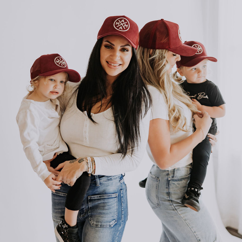 Two women in Mama X™ Brand Maroon Suede Mama X Trucker hats and white tops hold two children, all wearing matching red caps. The smiling group poses together against a white background.
