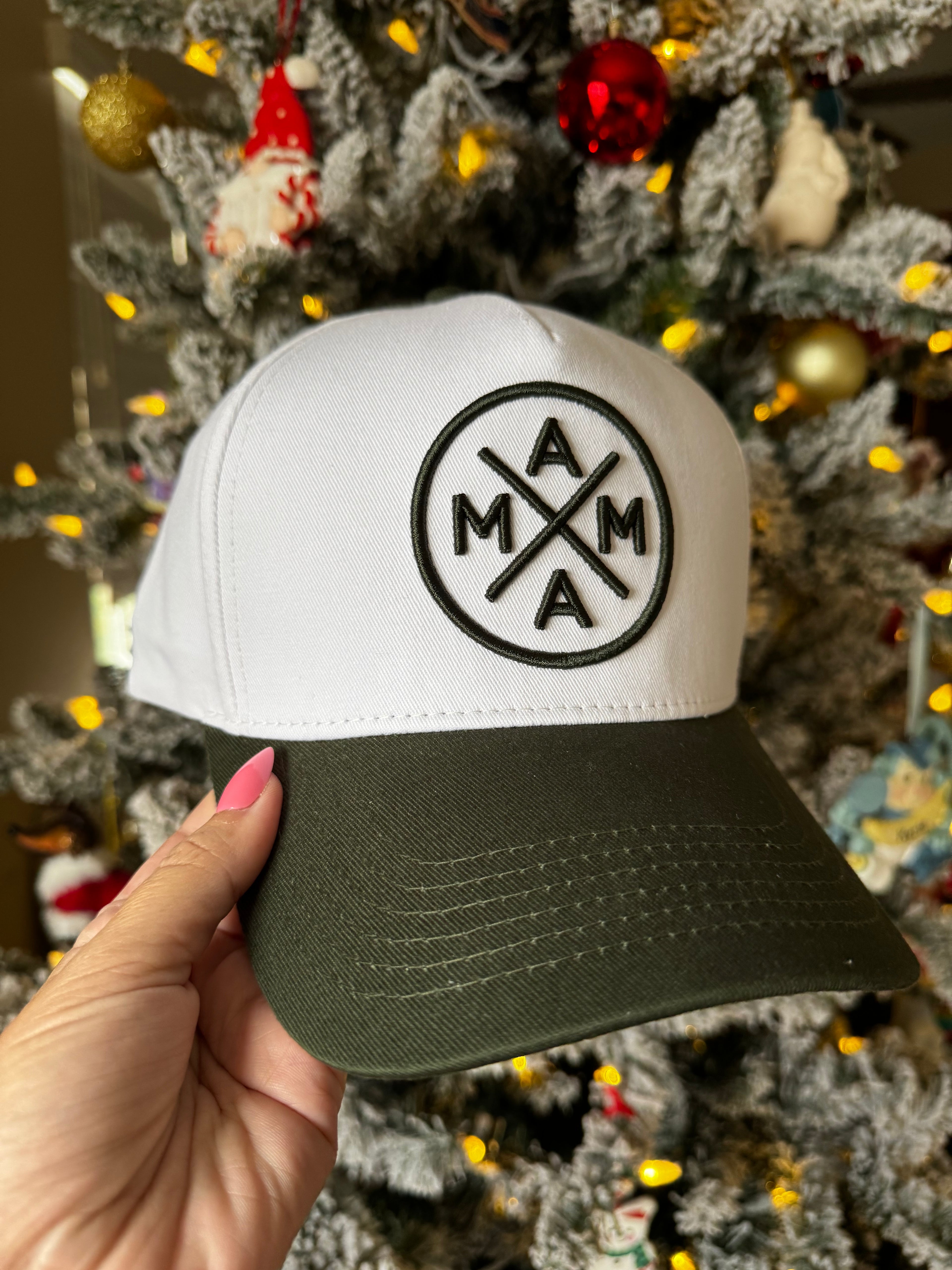 In front of a Christmas tree adorned with lights and ornaments, a hand displays a Dark Green Mama X™ Baseball Cap from the Mama X™ Brand. The cap features a circular logo with "MAMA" arranged in a cross pattern.