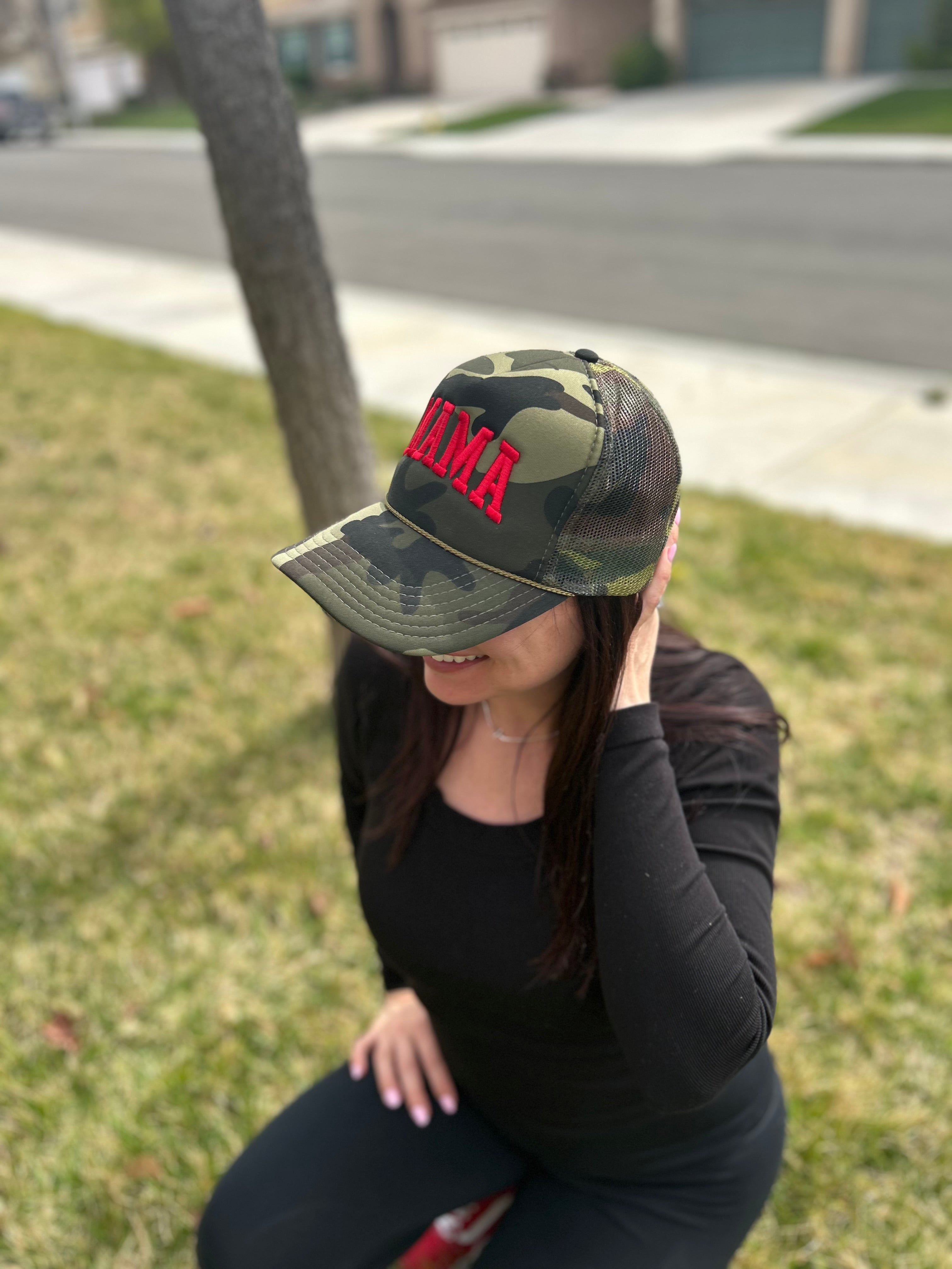 A woman in a black long-sleeve top and pants sits on grass, smiling and touching her hair, wearing the Mama X™ Brand Camo/Red Foam Trucker cap. A street and houses are visible in the background.