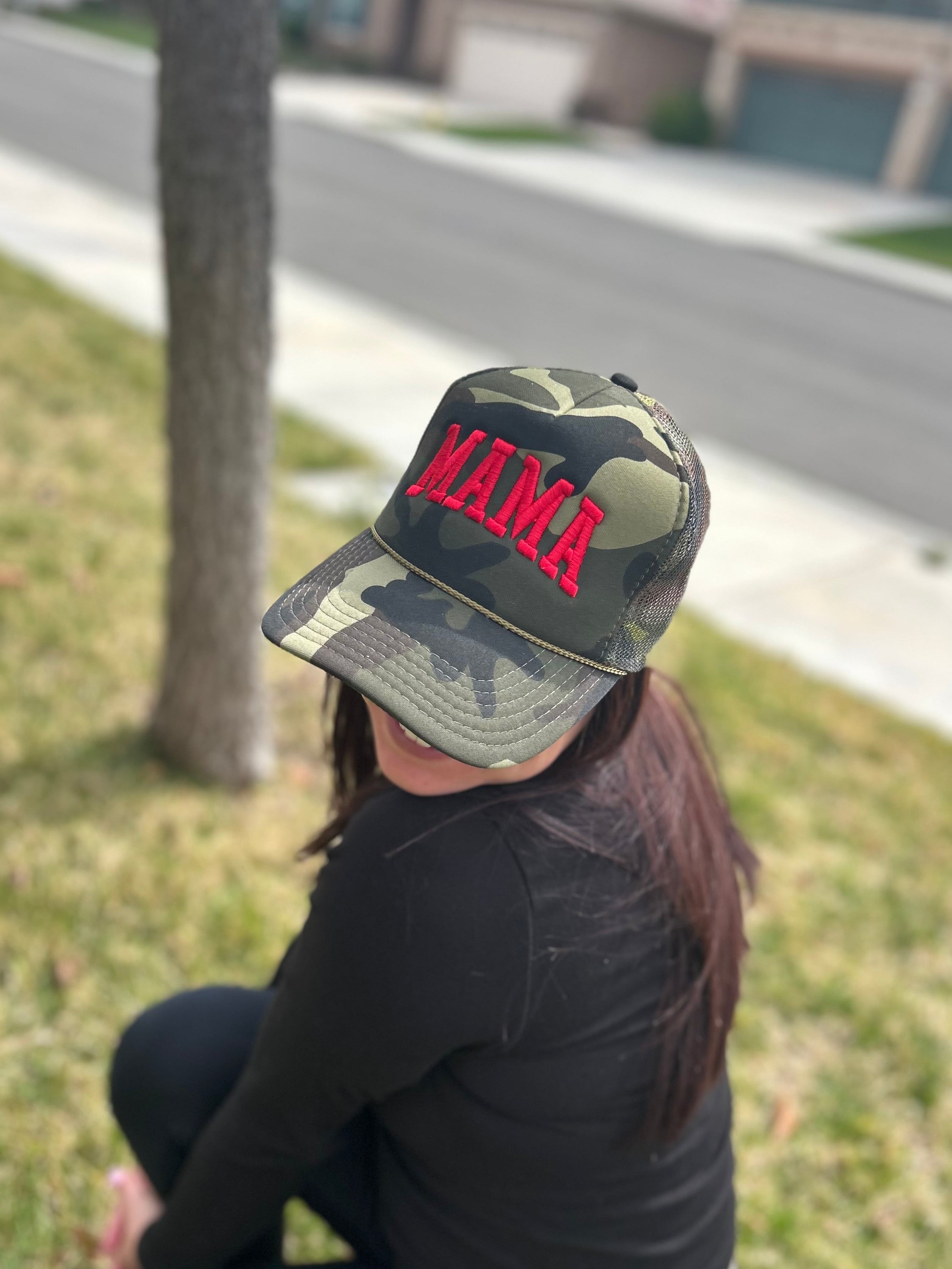 A person wears a Mama X™ Brand Camo/Red Foam Trucker cap with "MAMA" in red, sitting on the grass by a sidewalk and tree, blurred houses in the background.