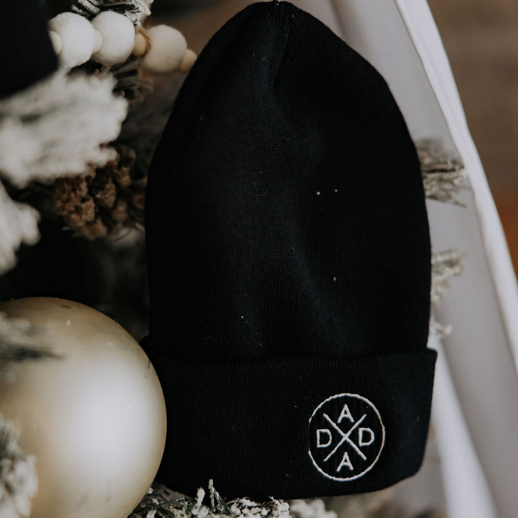 The Dada Cuffed Beanie by Mama X™ Brand, featuring a circular white "DADA" logo, is shown on a snow-dusted Christmas tree with festive accents—making it the perfect holiday gift for Dada.