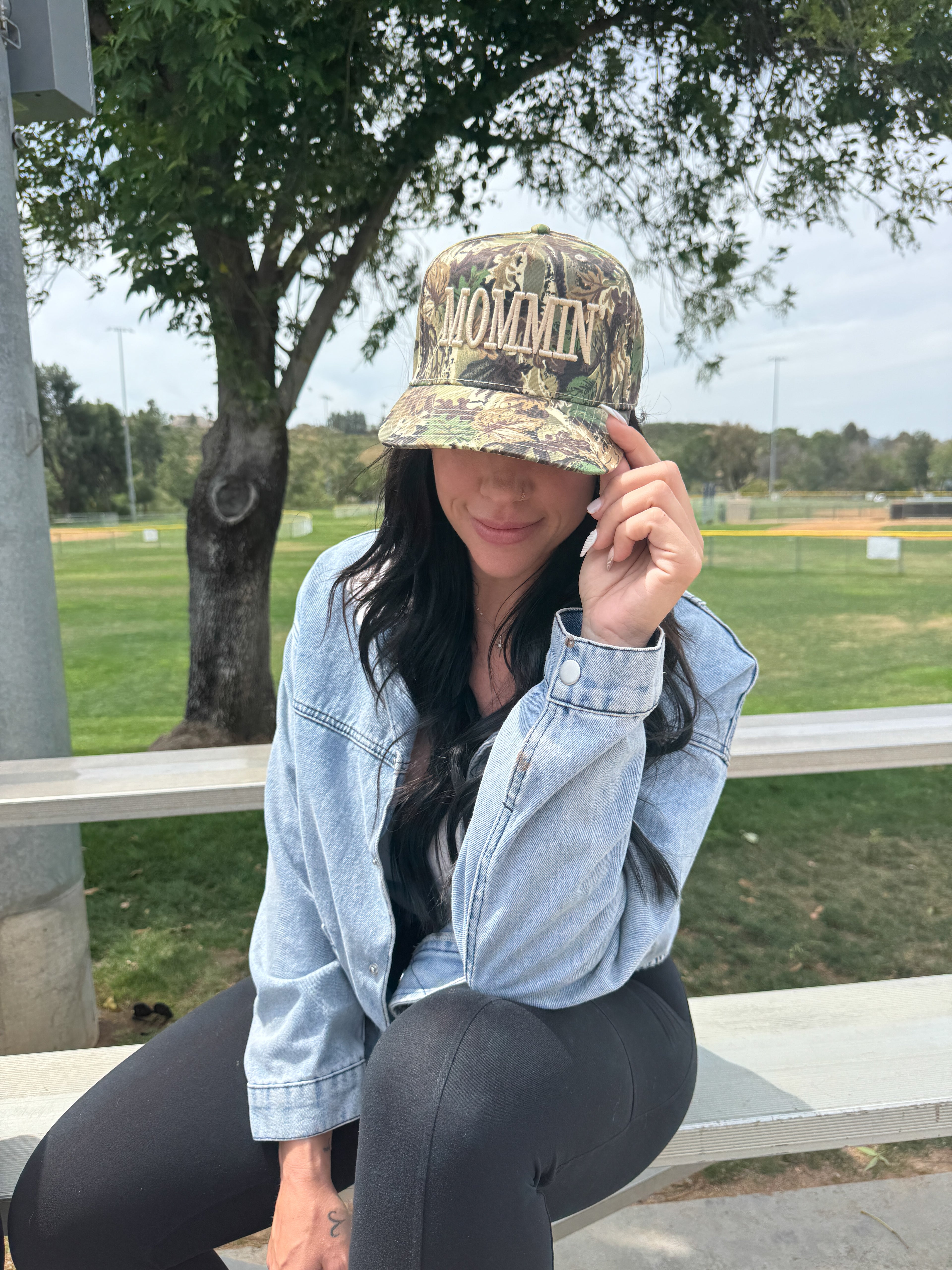 A woman sits on an outdoor bench wearing black leggings, a denim jacket, and the Mama X™ Brand Camo Mommin’ cap. She holds the brim while looking down, with trees and a park visible in the background.