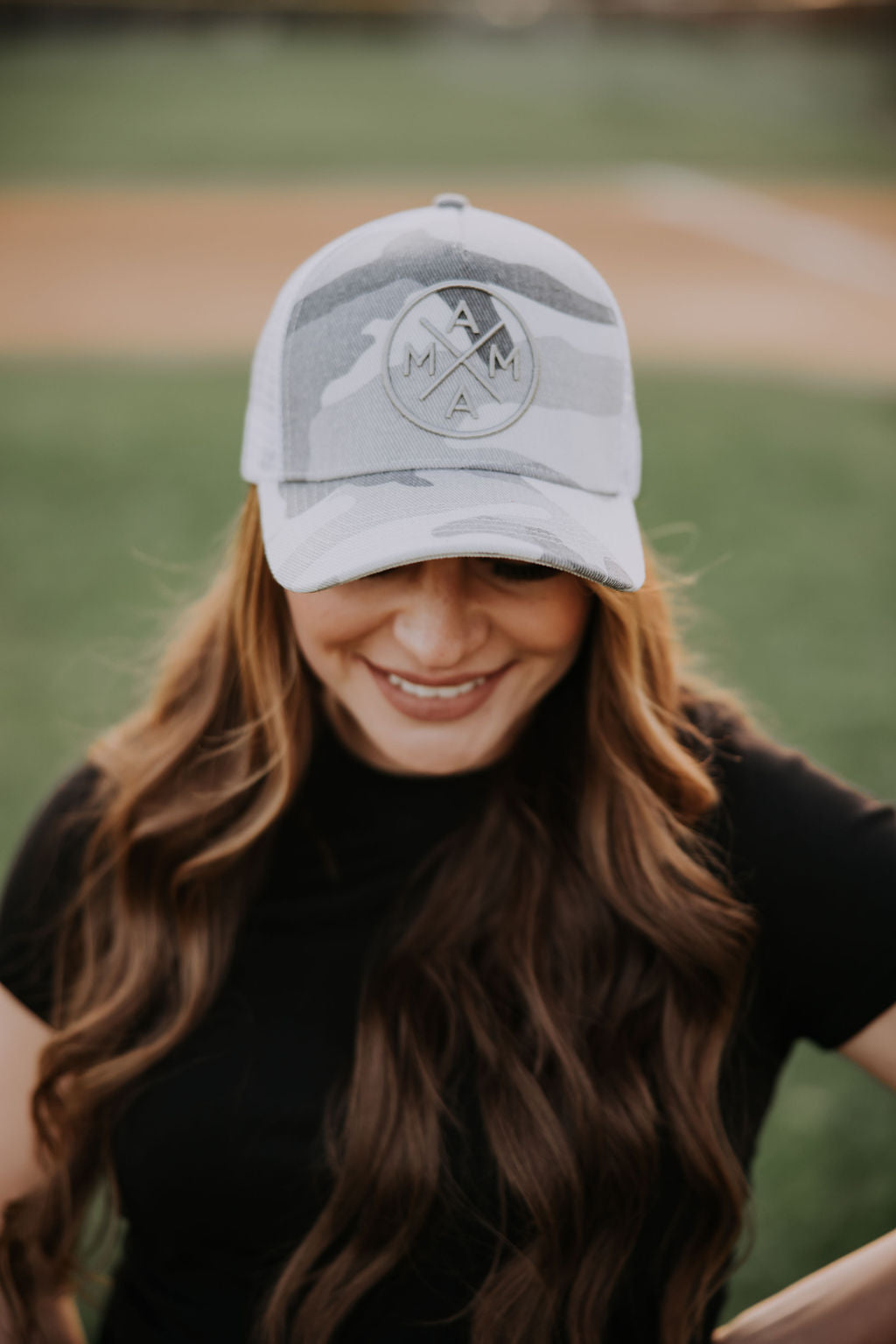 Wearing the White Camo Mama X™ Trucker Hat by Mama X™ Brand, featuring "MAMA" on the front, a person showcases their flowing brown hair over a black shirt. The adjustable snapback ensures mom pride shines against a blurred baseball field backdrop.