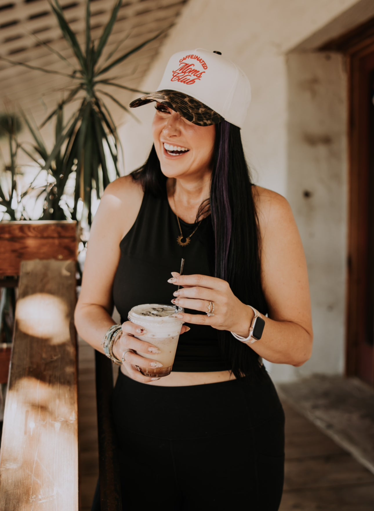 A woman with long dark hair smiles while holding a cold drink, wearing the Mama X™ Brand Caffeine Club Trucker cap. She stands indoors near a plant and wooden railing, dressed in a black sleeveless top and black pants.