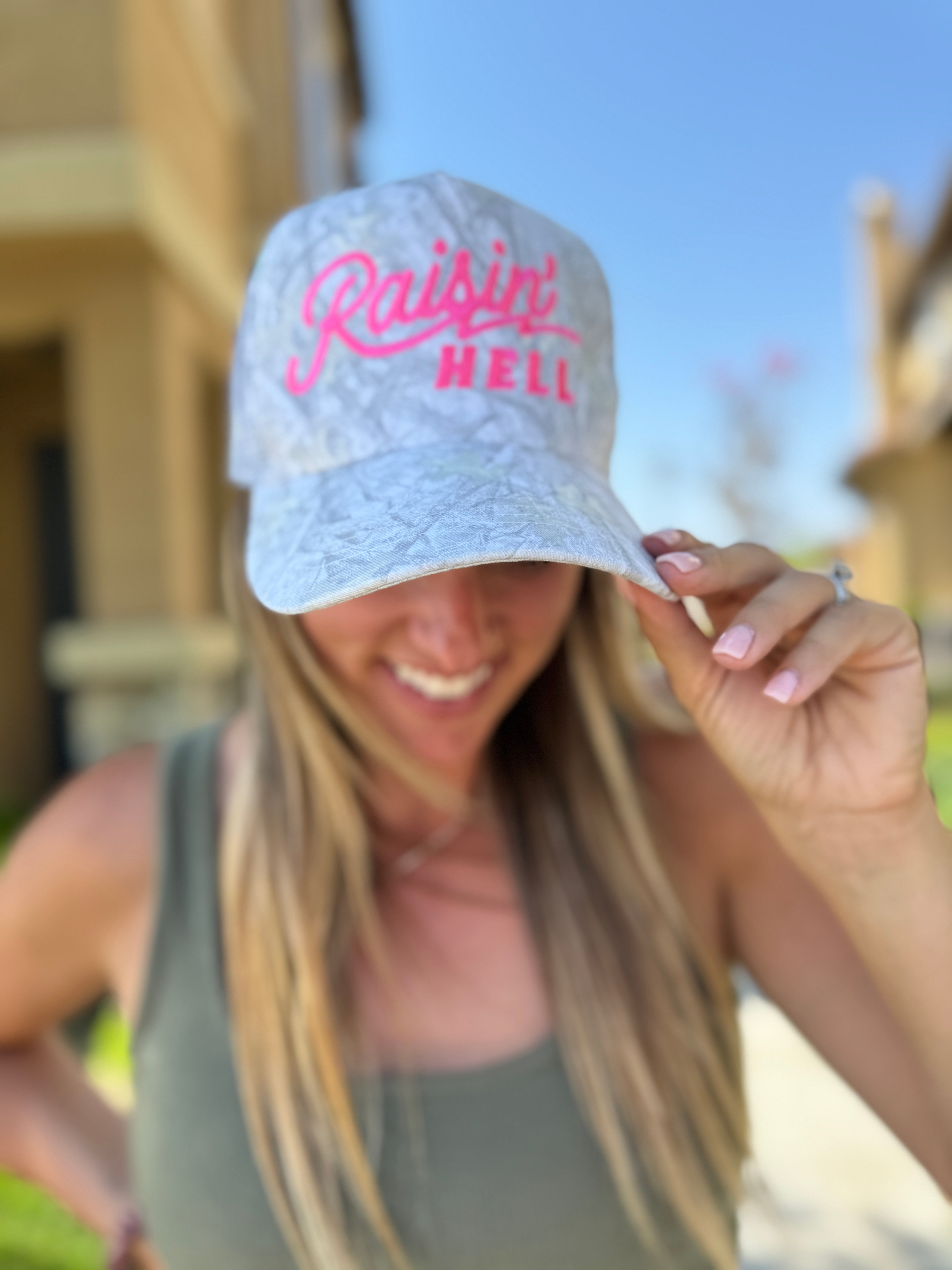 A smiling woman outdoors in a tank top shows off the Mama X™ Brand “Raisin’ Hell” Camo Trucker cap, playfully embodying motherhood spirit. The blurred background features sunlit buildings and greenery.
