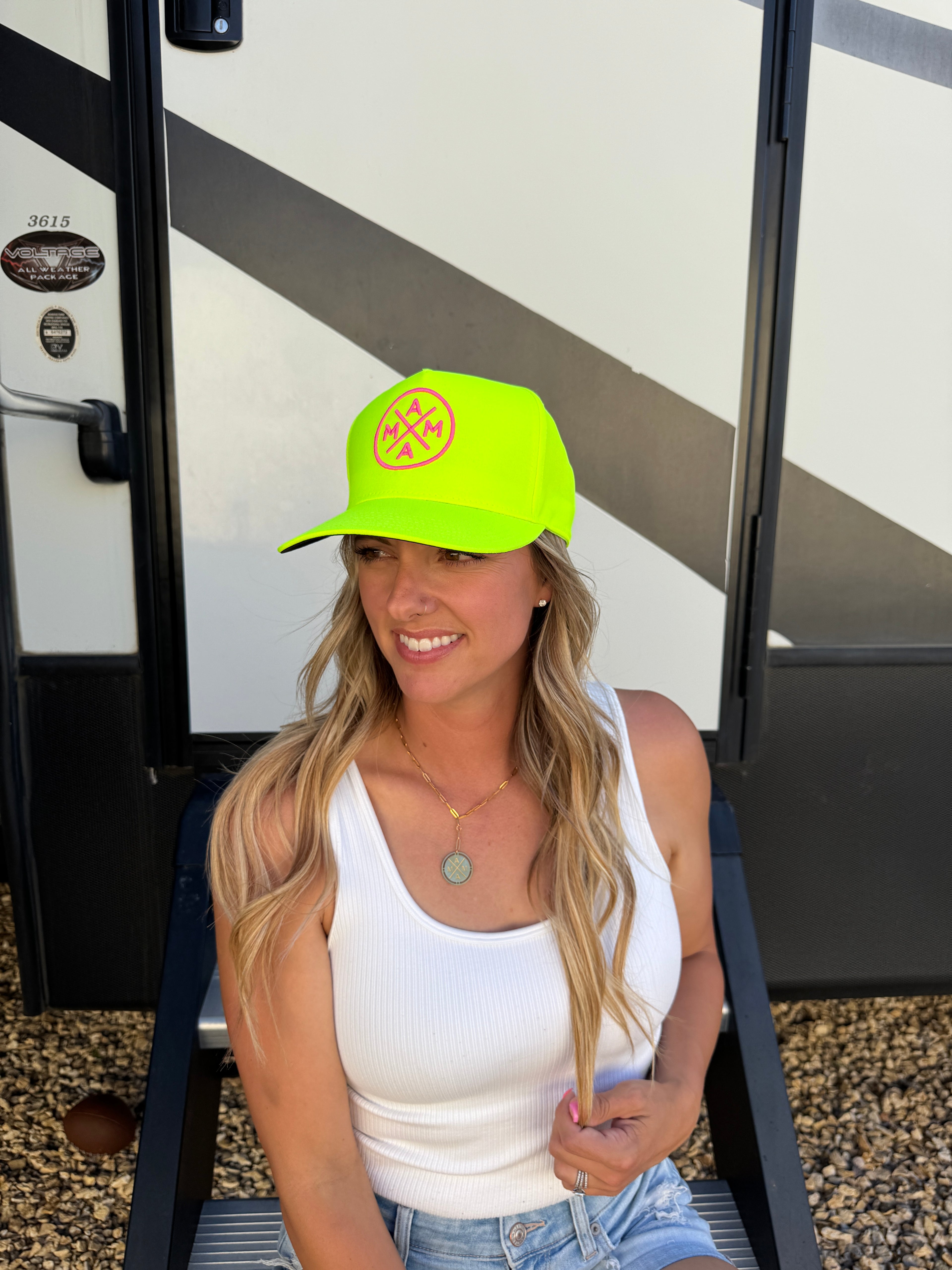 A woman with long blonde hair sits on steps in front of an RV, smiling and looking to the side. She wears a white tank top, jeans, and the Neon Yellow Mama X Trucker by Mama X™ Brand, featuring an adjustable snapback.