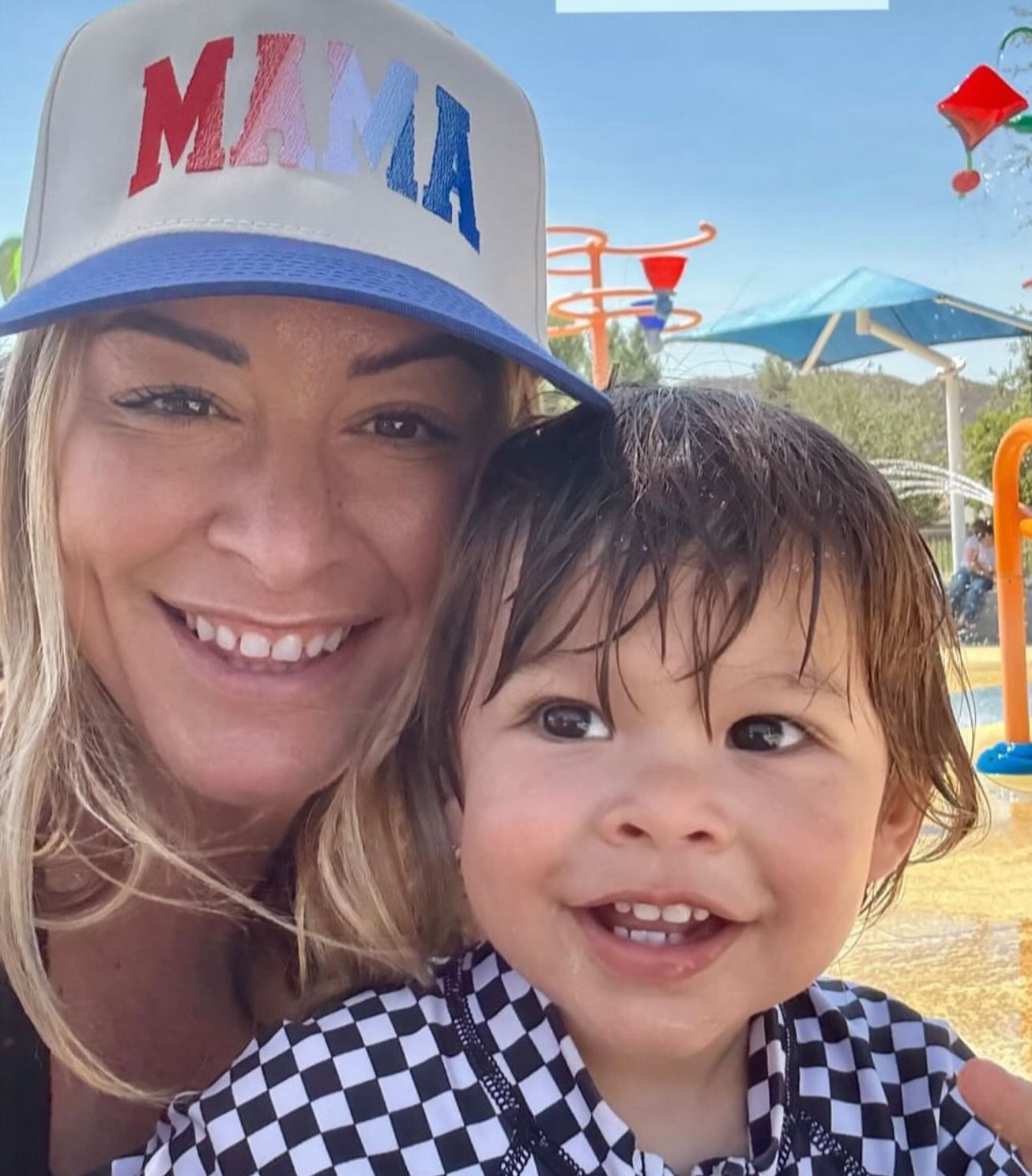 A smiling woman in a Red, White & Blue Mama Hat by Mama X™ Brand poses with a young child in a checkered outfit at a sunny outdoor splash park, with colorful playground equipment behind them.