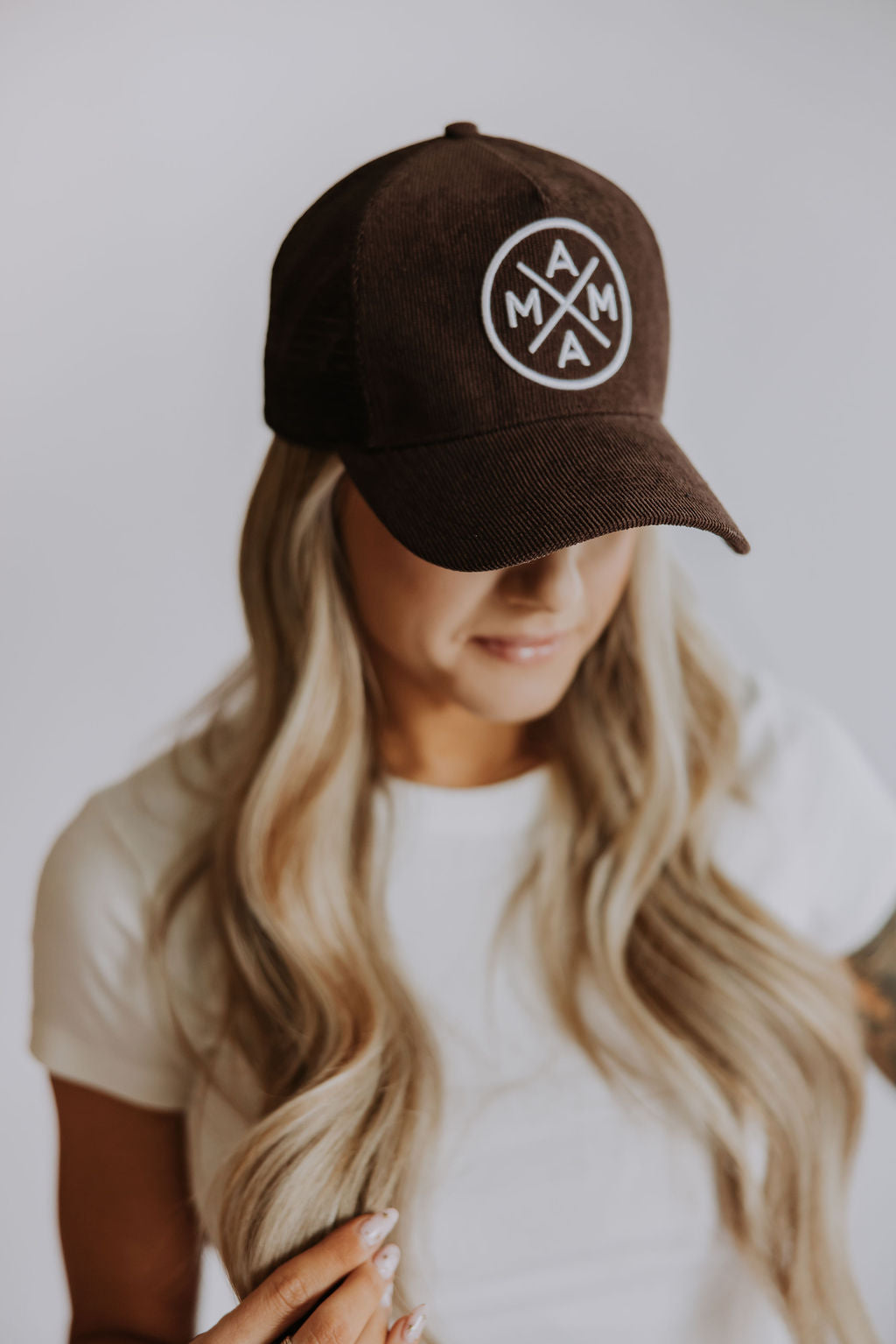 A woman with long, wavy blonde hair wears a white t-shirt and the Brown Mama X Corduroy Trucker by Mama X™ Brand, featuring a circular "MAMA" logo. She looks downward, partially obscuring her face against a plain, light background.