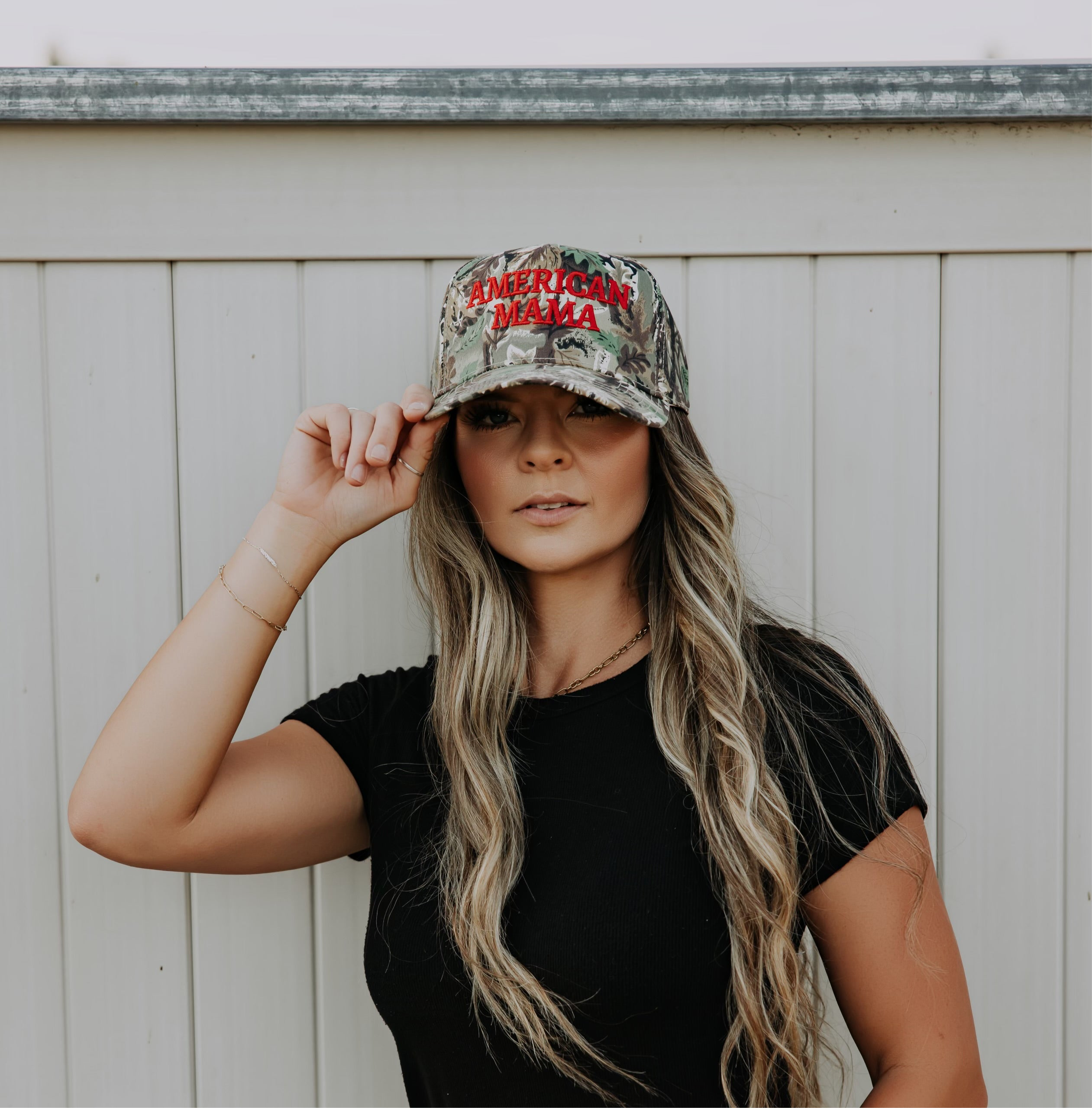 A woman with long, wavy hair wears a black t-shirt and the Mama X™ Brand American Mama Hat—a camo cap with “AMERICAN MAMA” on the front—while standing by a white wooden fence and holding the brim of her hat.