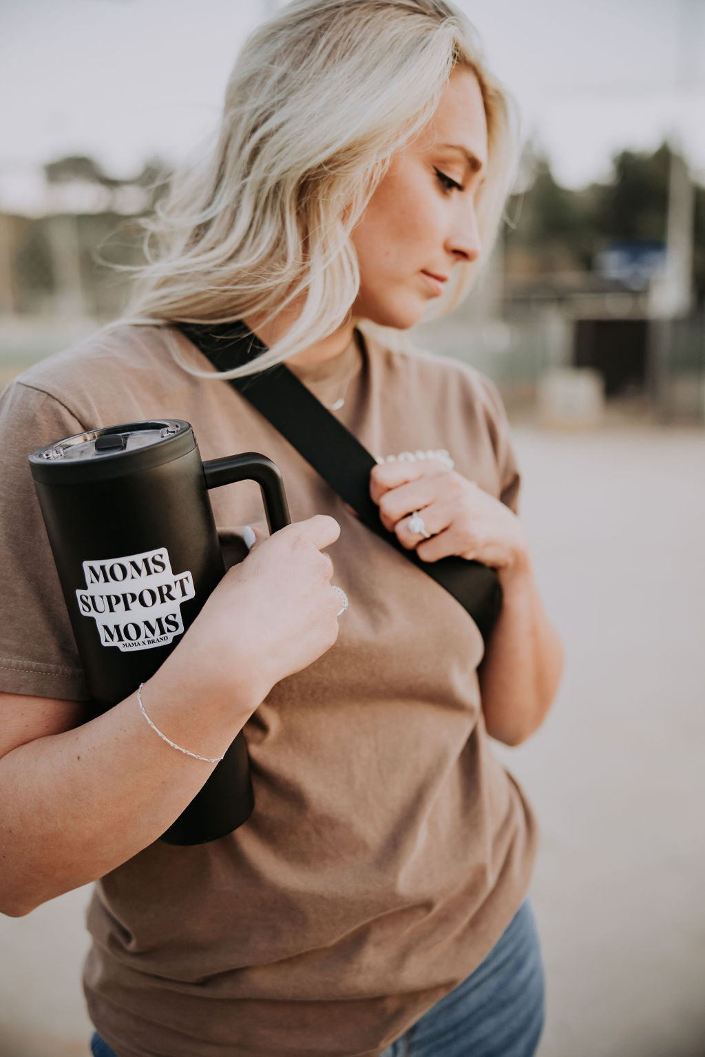 A blonde woman with a black tumbler displays a "Moms Support Moms" sticker from Mama X™ Brand. She sports a brown t-shirt and a black shoulder bag, ideal for an outing, set against a blurred outdoor scene that highlights the sticker's supportive message in daily life.
