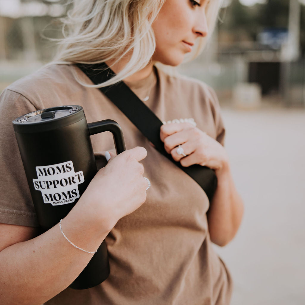 A blonde woman with a black tumbler displays a "Moms Support Moms" sticker from Mama X™ Brand. She sports a brown t-shirt and a black shoulder bag, ideal for an outing, set against a blurred outdoor scene that highlights the sticker's supportive message in daily life.