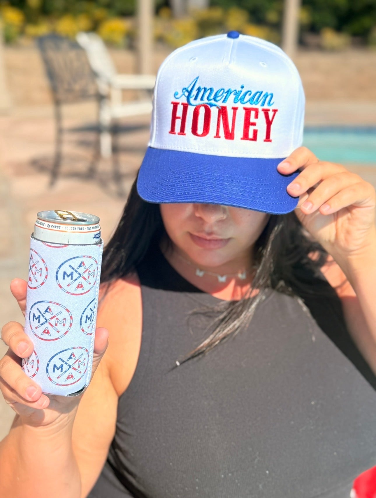 A woman in a black sleeveless top holds a canned drink with the Mama X™ Brand "Mama X American Flag Can Cooler" and tips a blue and white "American Honey" hat over her eyes. She stands outdoors near a pool under sunlight.