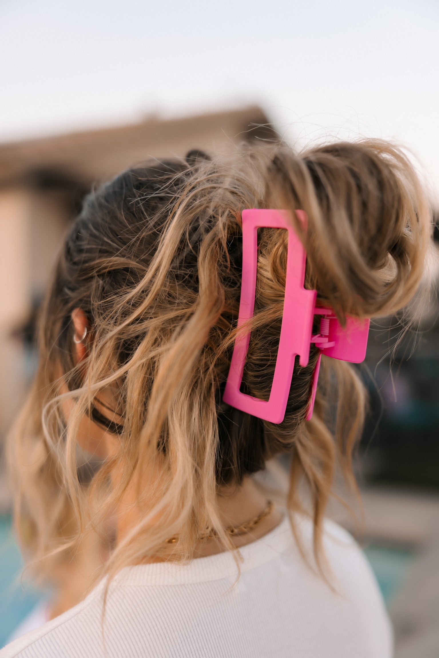 A woman with wavy, thick light brown hair wears it in a messy bun, secured with the Mama X™ Brand Pink Claw Clip - XL. She is dressed in a white top and photographed from behind.