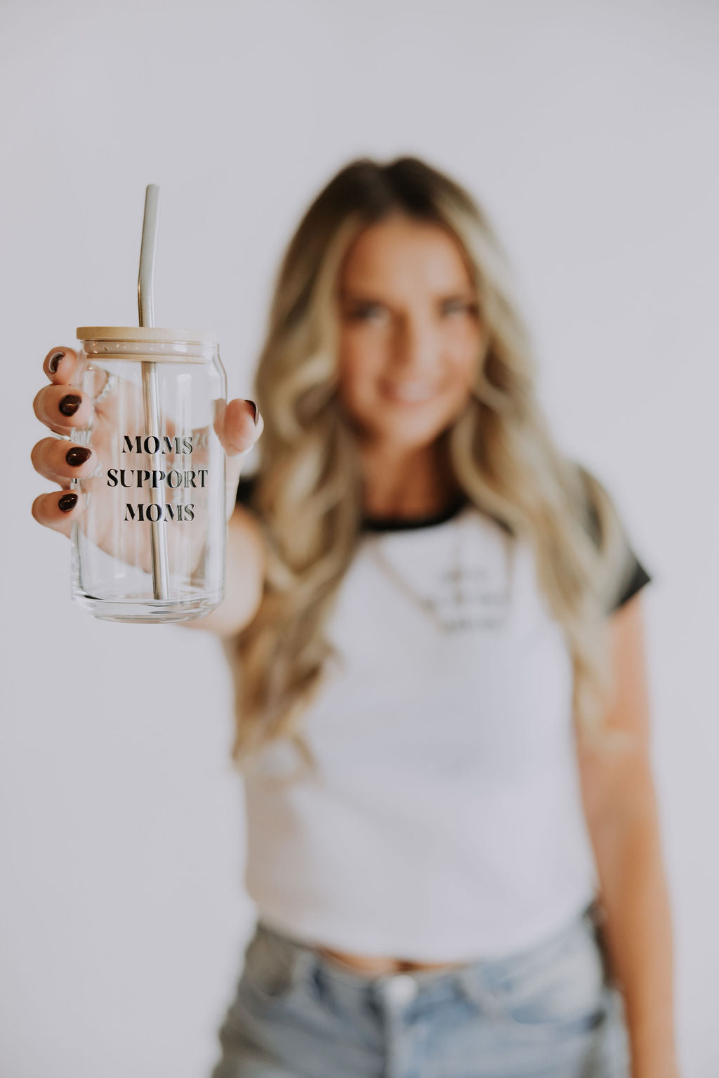 A woman with long blonde hair, in a black and white tee and jeans, holds a 16 oz Mama X™ Brand "Moms Support Moms Glass" mug with straw. The background is plain and light.