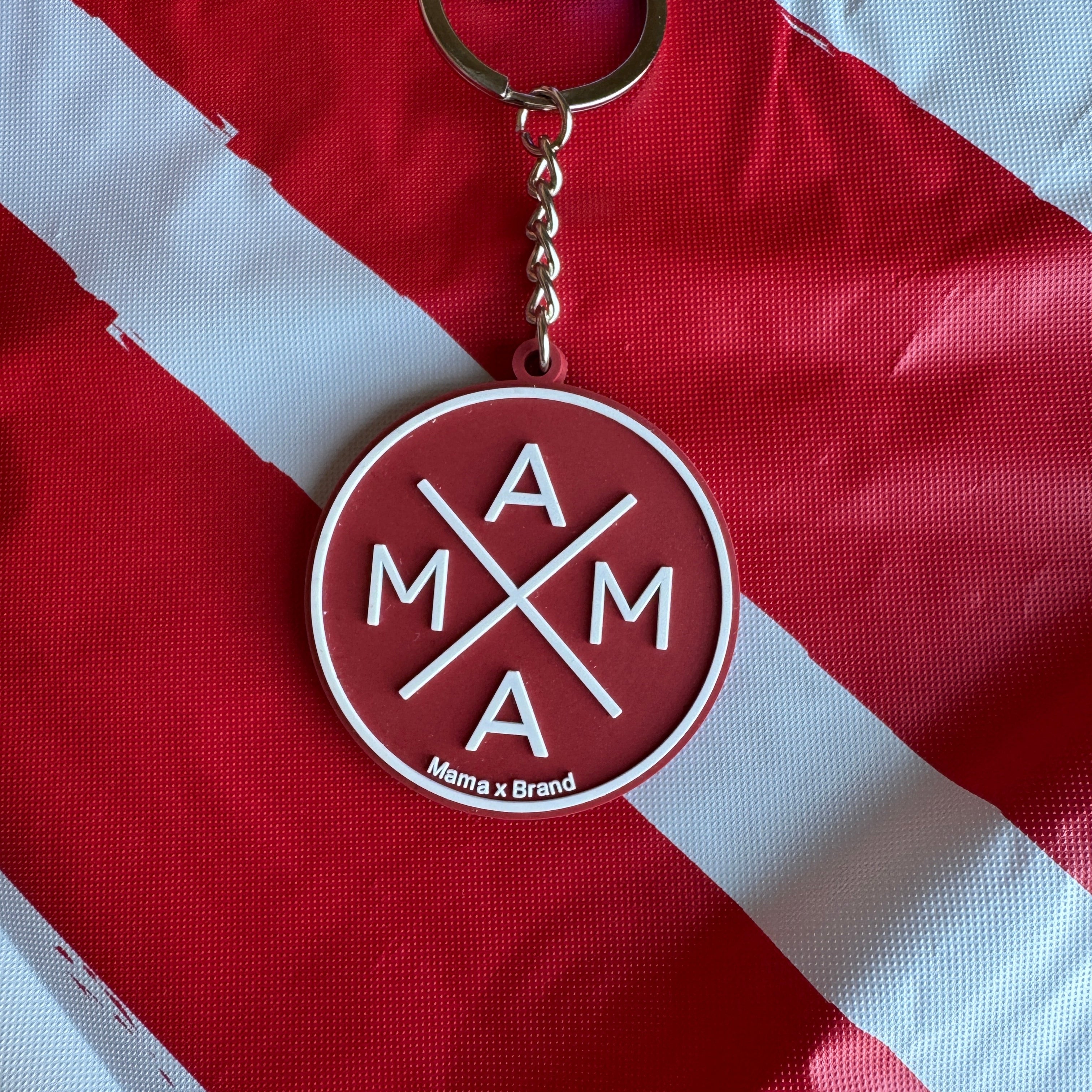 The Red Mama X Keychain by Mama X™ Brand, featuring "MAMA" and crossed lines on a red background, rests on red and white striped fabric.