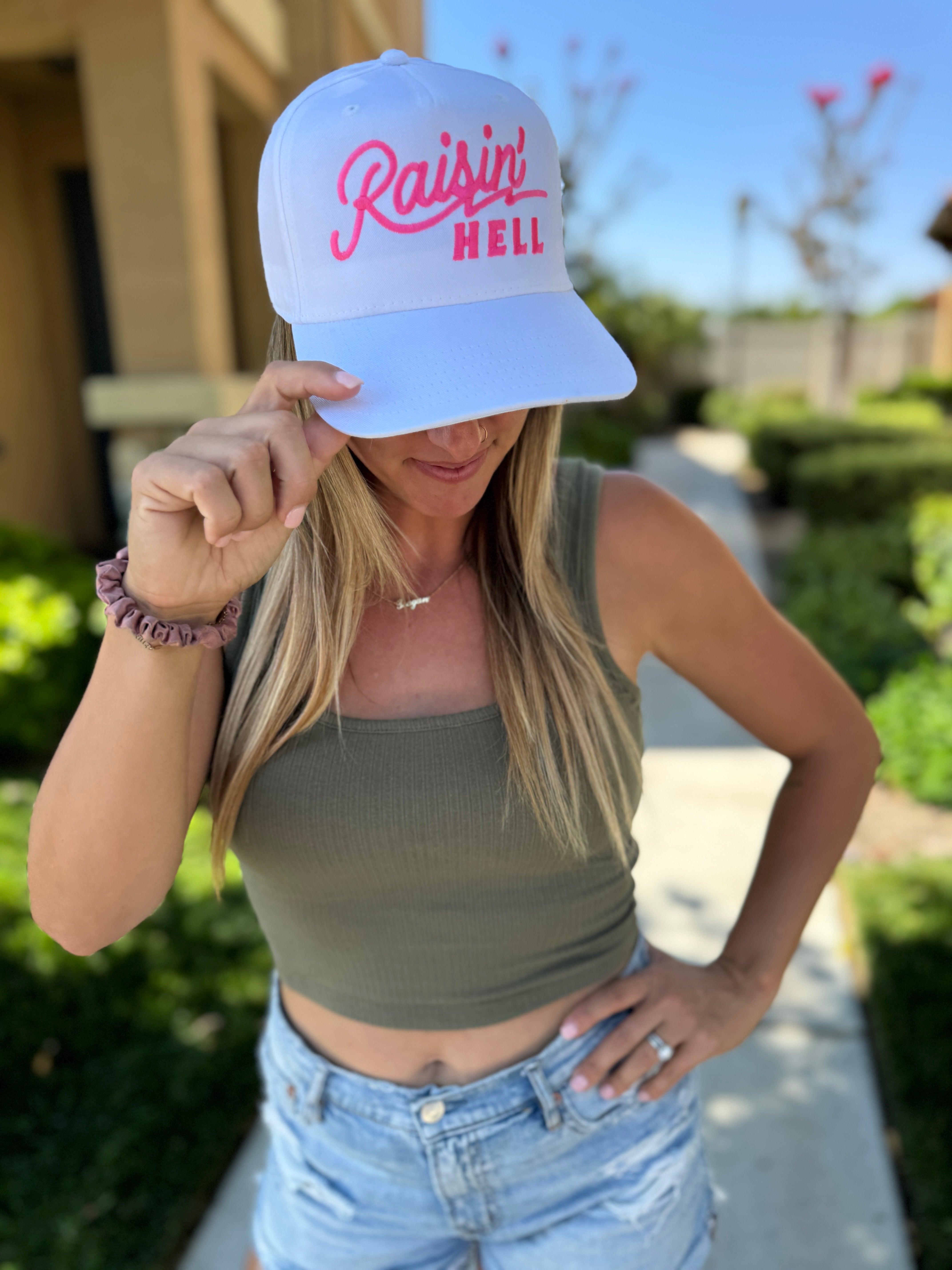 A woman in a green tank top and denim shorts stands outside, playfully holding her Mama X™ Brand Raisin’ Hell White Trucker cap—perfectly capturing that raisin' hell or raisin' kids energy.