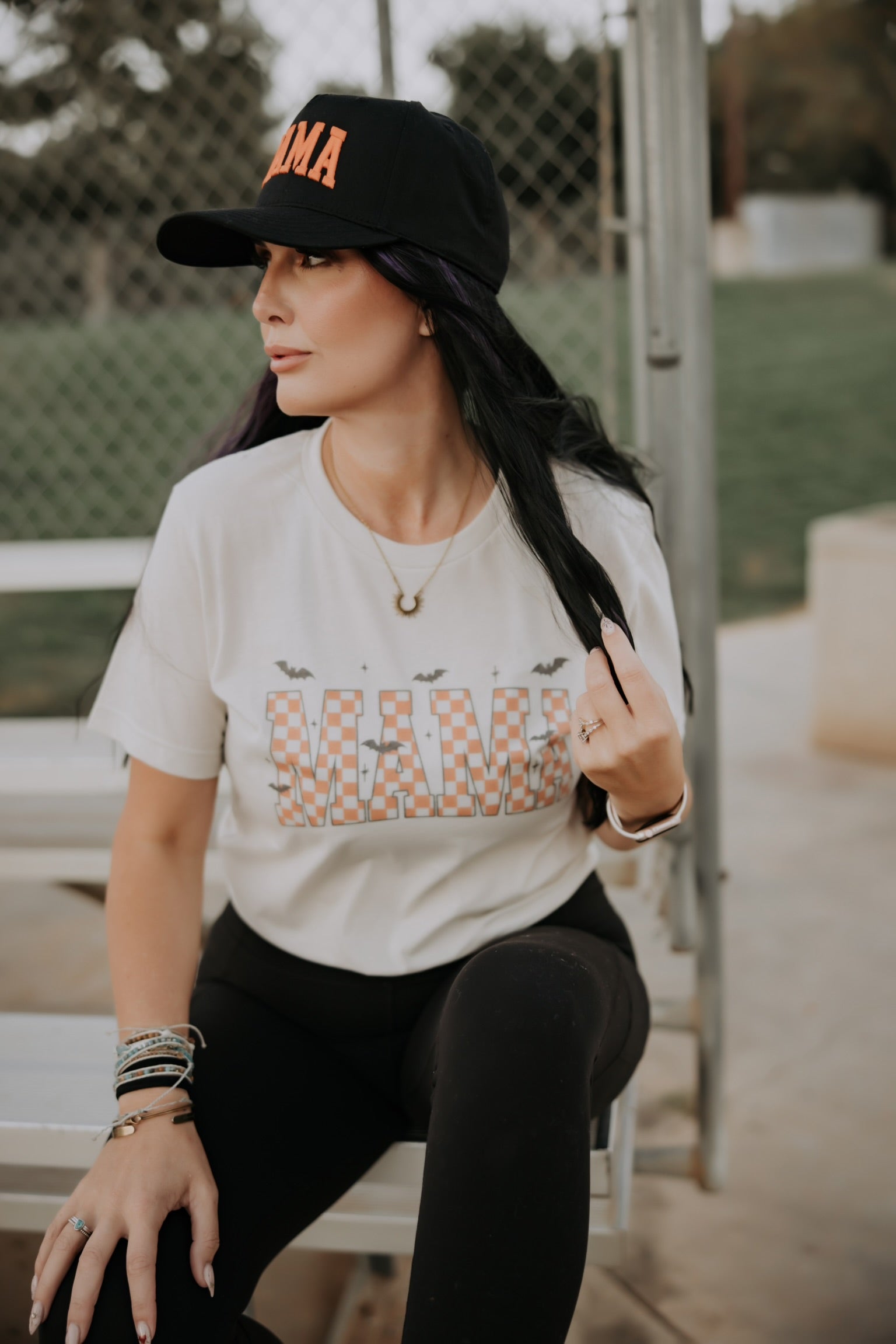 Woman with long dark hair wears a black cap and the Mama X™ Brand Mama Halloween Tshirt with black leggings and bracelets, sitting on an outdoor bench by a chain-link fence, looking to the side.