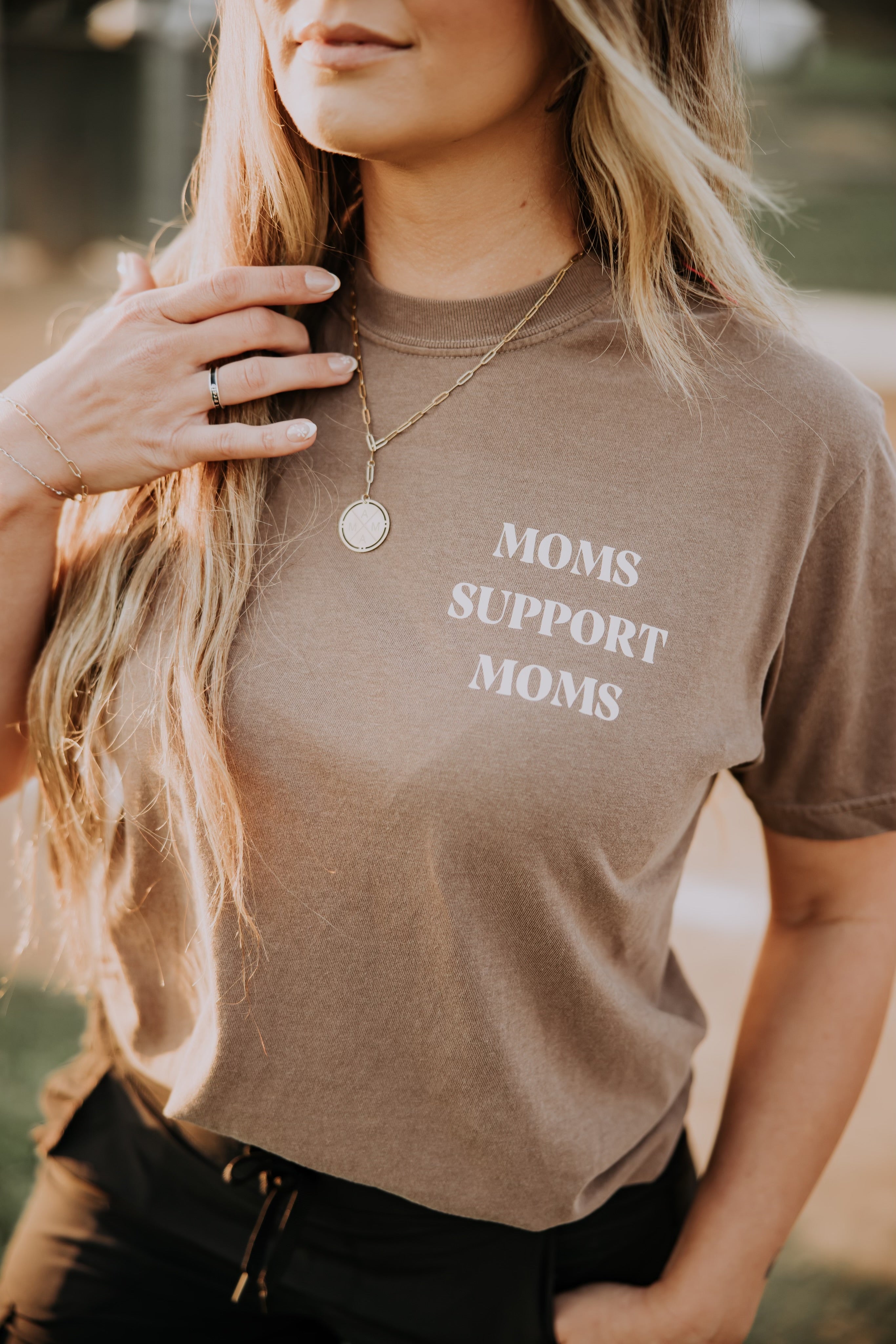A person with long, blonde hair wears a garment-dyed brown "Moms Support Moms" T-shirt from Mama X™ Brand, featuring a necklace and a ring on their left hand. The blurred background suggests an outdoor setting, capturing the essence of a supportive mama.