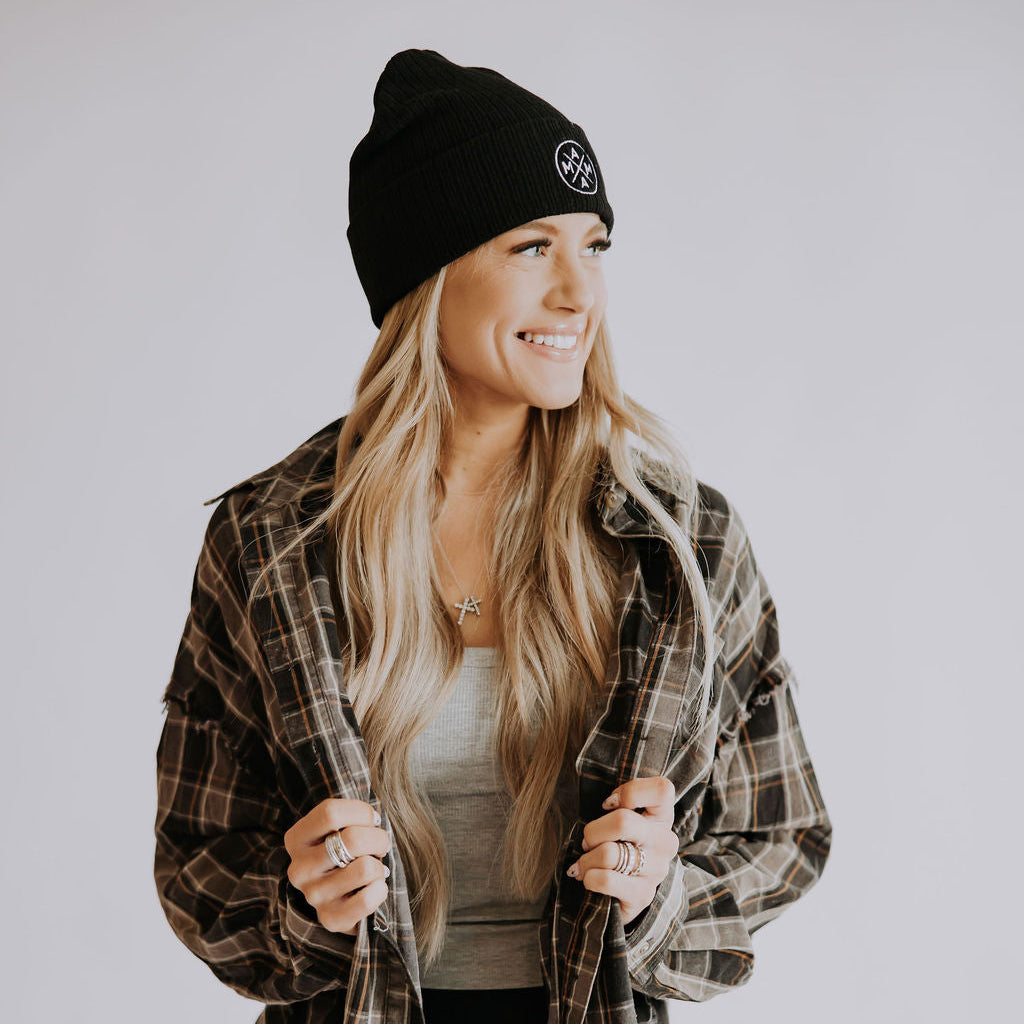 A woman with long blonde hair smiles and looks to the side against a plain, light background, wearing the Mama Cuffed Beanie by Mama X™ Brand with a plaid flannel over a gray top and black pants.