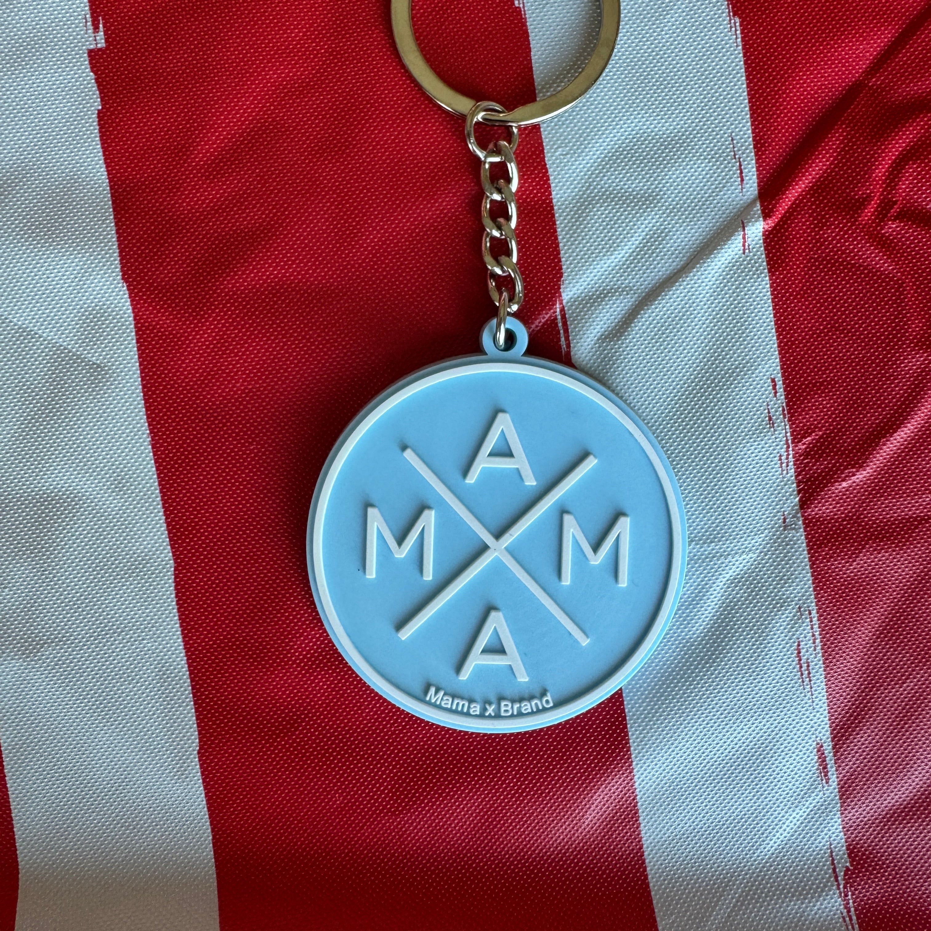 The Blue Mama X Keychain by Mama X™ Brand features "MAMA" in a cross pattern on a round blue charm with a gold chain, displayed on red and white striped fabric.