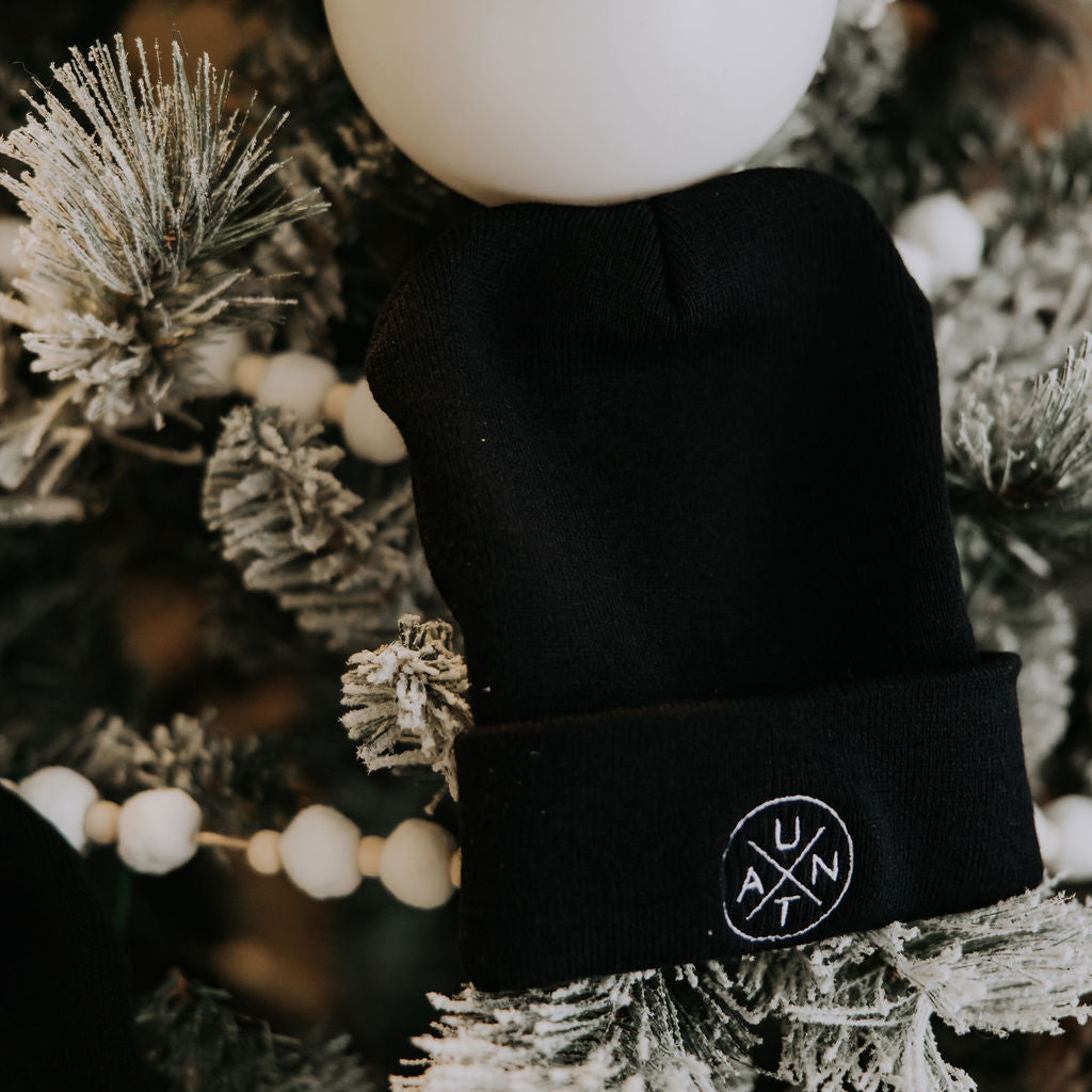 The Mama X™ Brand Aunt Cuffed Beanie—a cozy black knit hat with a white round logo—hangs on a snow-dusted holiday tree, making it the perfect gift for your aunt this Christmas season.