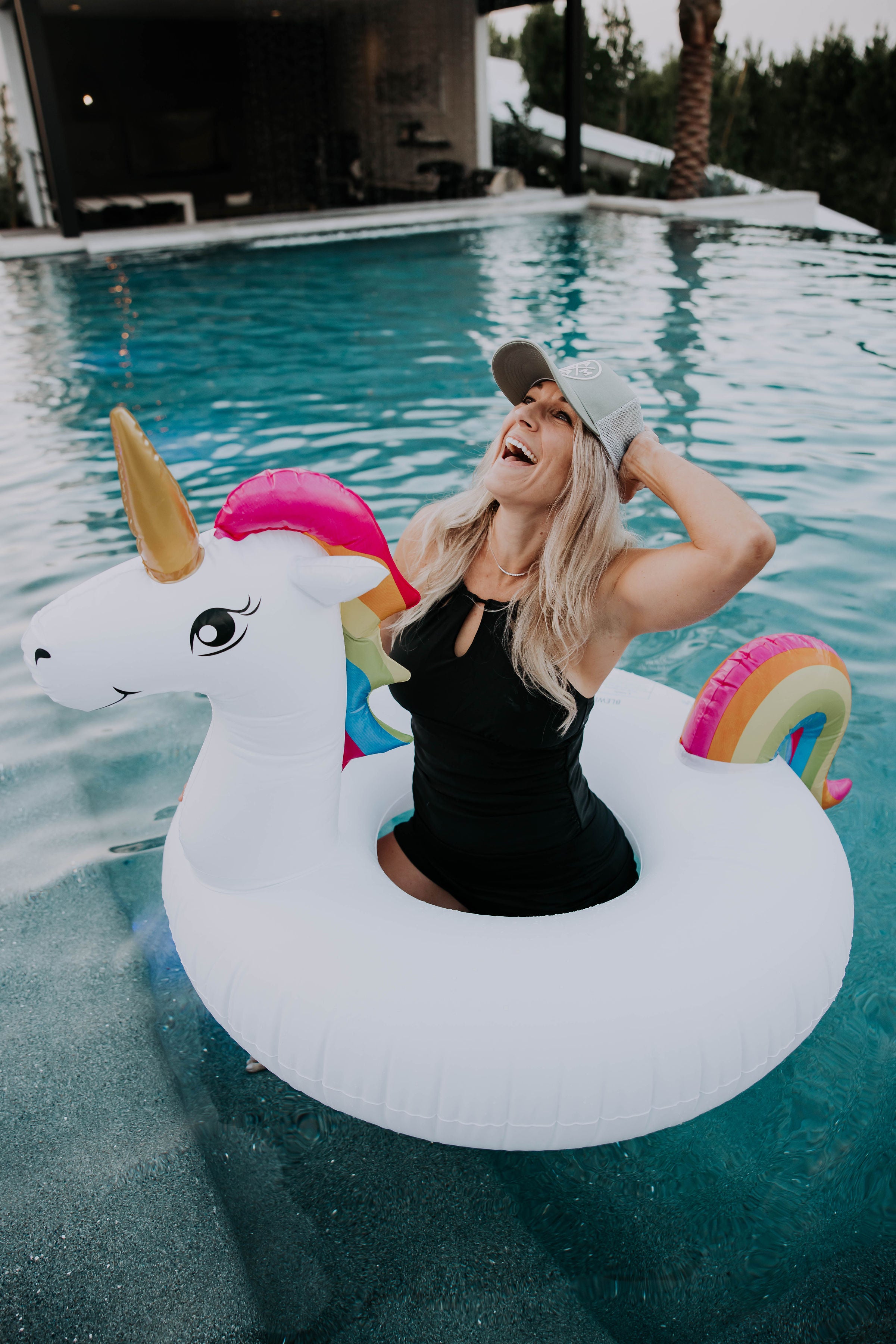 A woman with long blonde hair, wearing a black swimsuit and a Mama X™ Premium Canvas Trucker Hat - Sage from Mama X™ Brand, is laughing cheerfully while sitting on a white unicorn pool float in a swimming pool. The unicorn float has a rainbow mane and tail. Trees and a building are visible in the background.