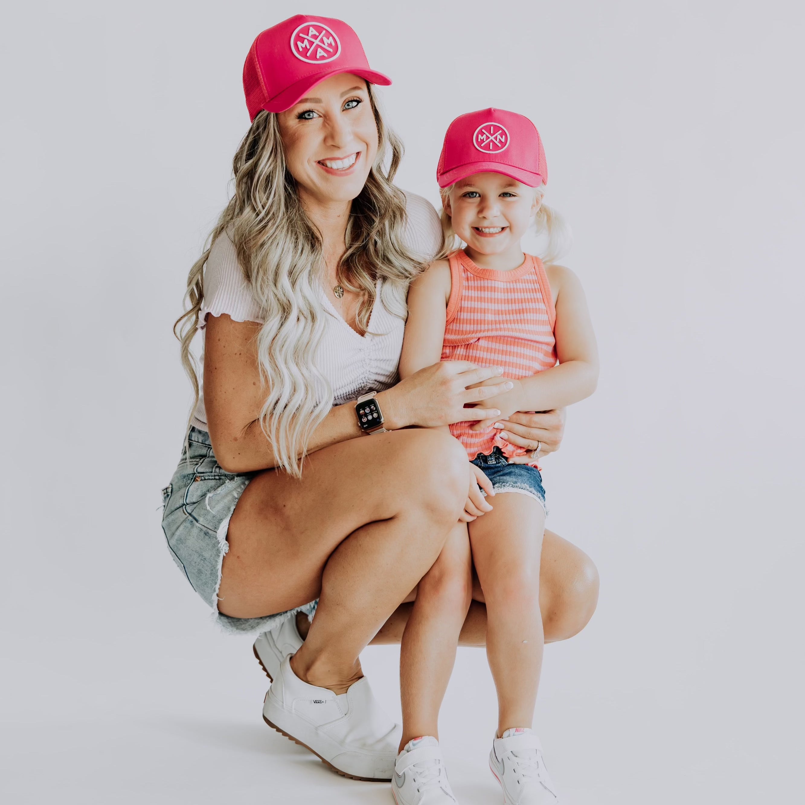 A woman with long, wavy hair and a young girl with pigtails smile at the camera. Both are wearing matching Mini X Premium Trucker Hats in hot pink from Mama X™ Brand, casual summer outfits, and white sneakers. They are positioned against a plain white background.