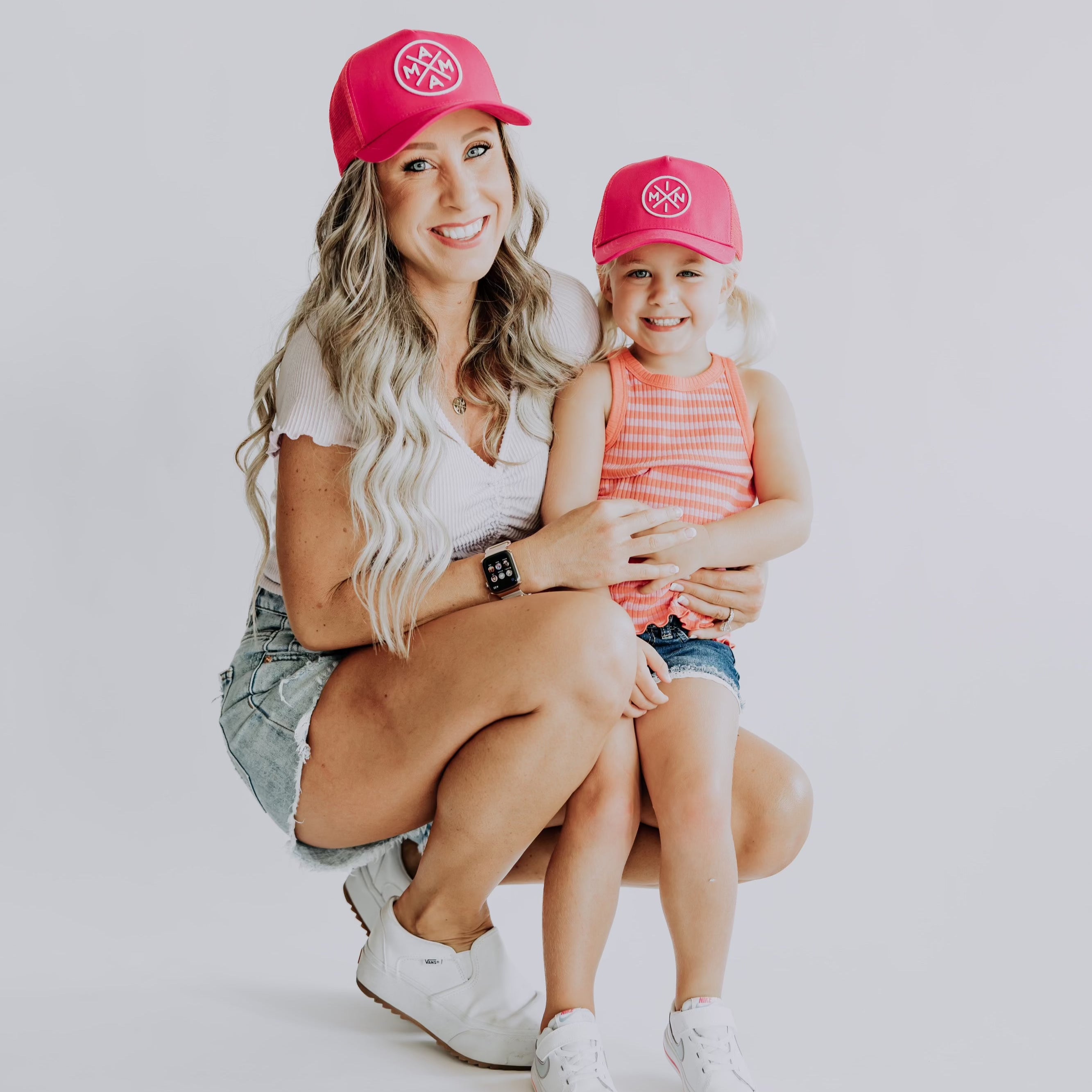 A woman and a young girl, both sporting matching hot pink Mama X™ Premium Canvas Trucker Hats by Mama X™ Brand, smile at the camera. The woman, with long wavy hair, is dressed in a white top, denim shorts, and sneakers. The child, with blonde pigtails, wears an orange tank top, denim shorts, and white sneakers. They pose against a plain background.