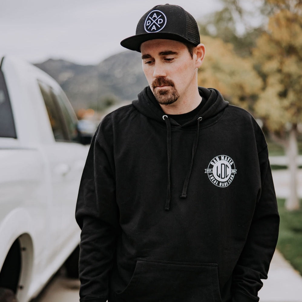 A man with a beard and mustache stands near a white truck. He is wearing a black cap and the Dad Mafia Hoodie from Mama X™ Brand, which is a heavy-weight hoodie made of ring-spun cotton featuring a logo on the front. The background showcases blurred trees and hills.