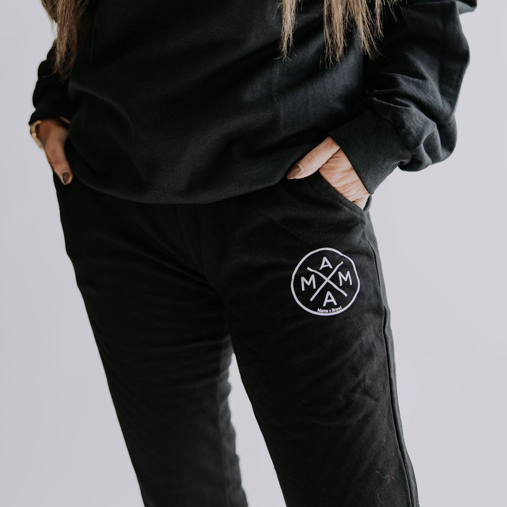 The individual is wearing a classic fit, cozy everyday ensemble that includes a black sweatshirt paired with Mama X™ Brand's Mama X™ Joggers. The joggers display a circular logo featuring the word "MAMA" in a cross-pattern design. Their hands are casually tucked into the pockets, set against a simple, light-colored background.