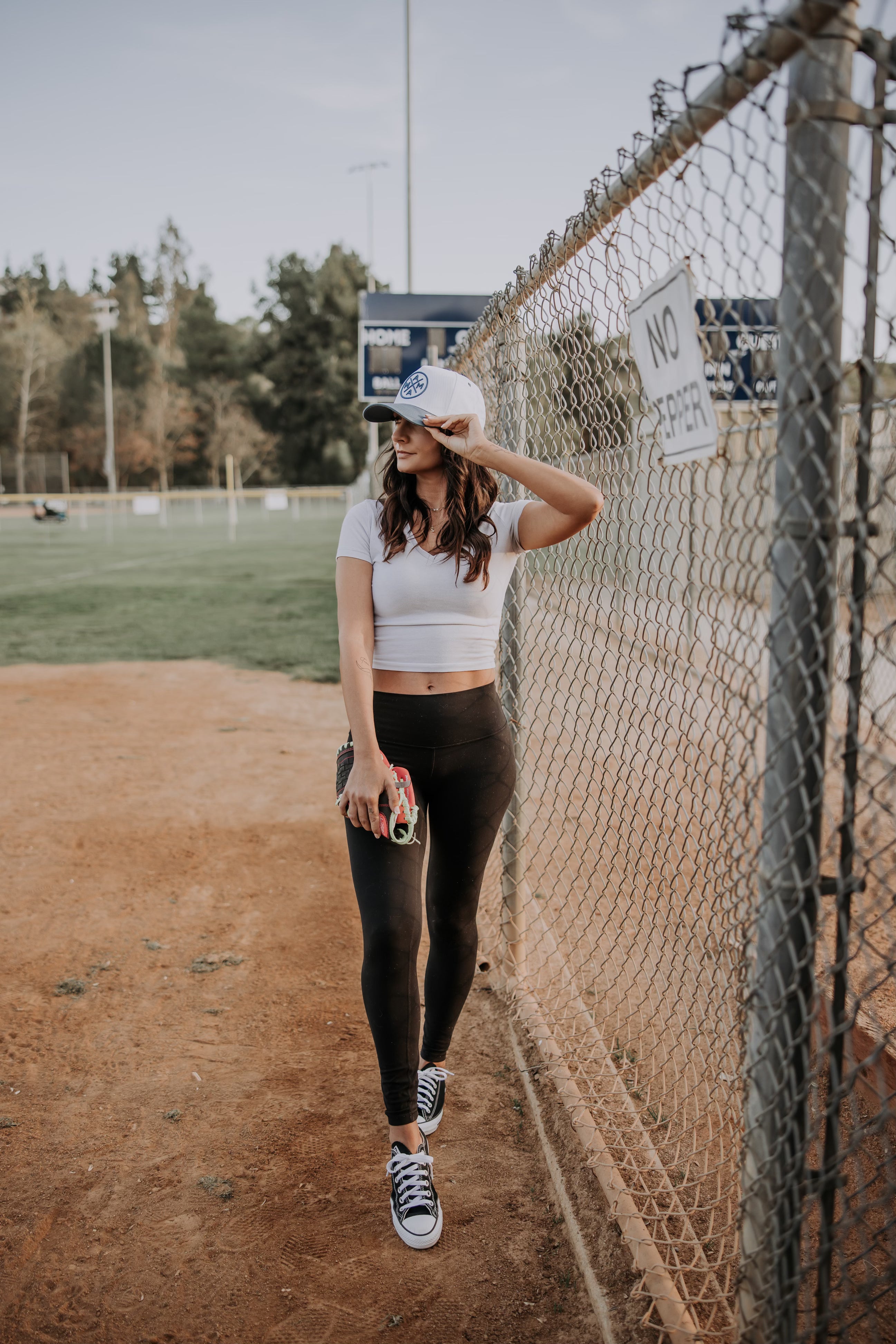 A woman stands by a chain-link fence on a sports field, wearing a white crop top, black leggings, sneakers, and the Blue Mama X™ Baseball Cap by Mama X™ Brand. She holds a can and gazes sideways with trees and a scoreboard in the background.