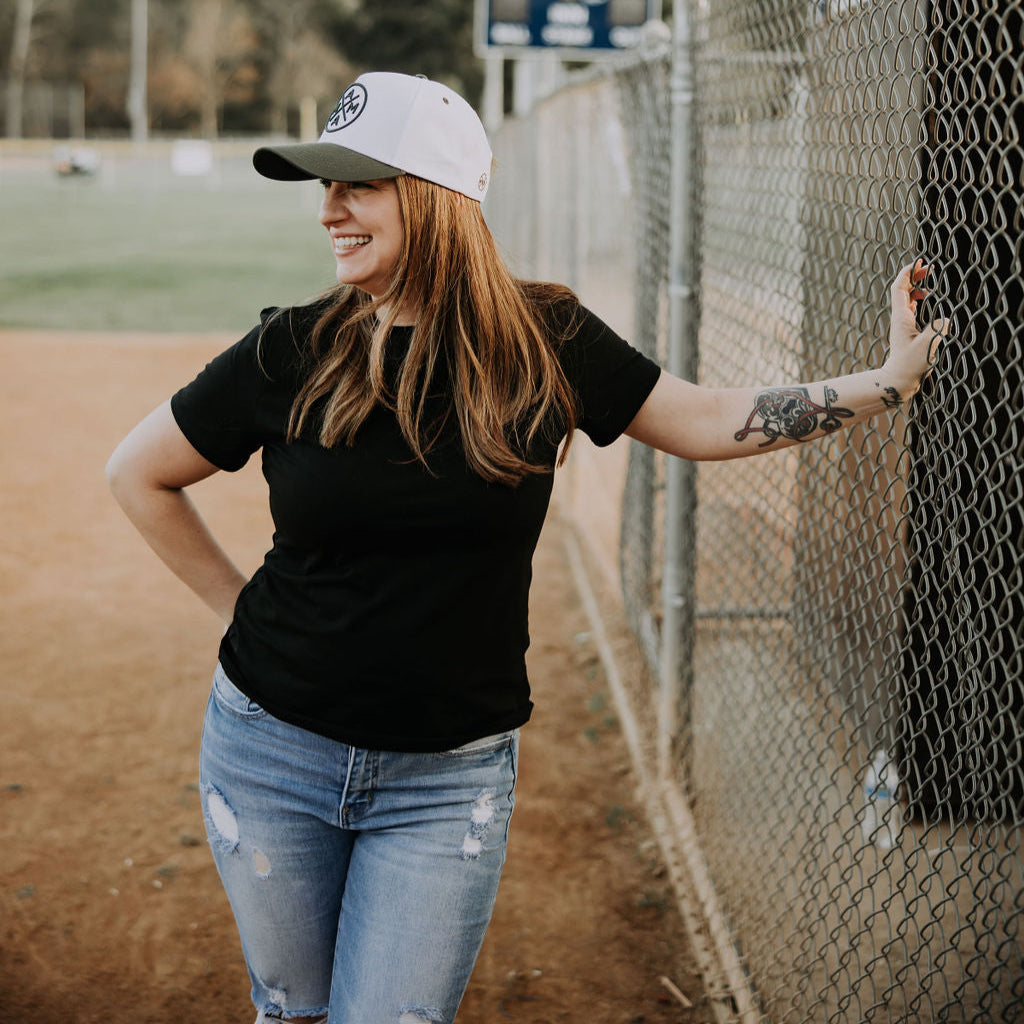 A person in a black shirt and jeans leans against a chain-link fence on a baseball field, sporting the best-seller Dark Green Mama X™ Baseball Cap. With long hair and a tattooed arm, they stand amidst trees and a scoreboard in the background, capturing an effortless holiday vibe.