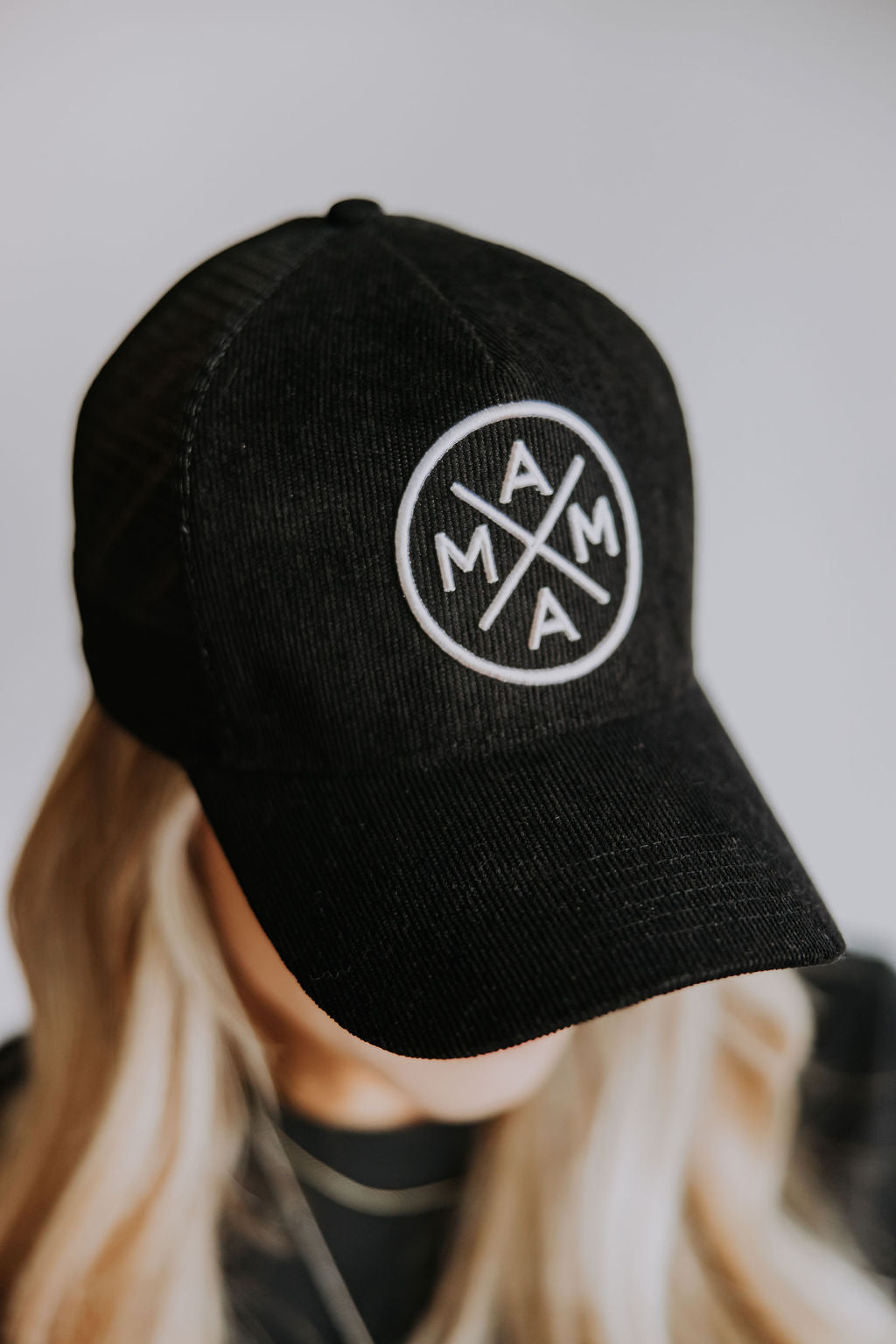 A person with long blonde hair wears the Black Corduroy Mama X Trucker by Mama X™ Brand, featuring a white circular "MAMA" logo on the front. The person’s face is mostly out of view, looking downward.