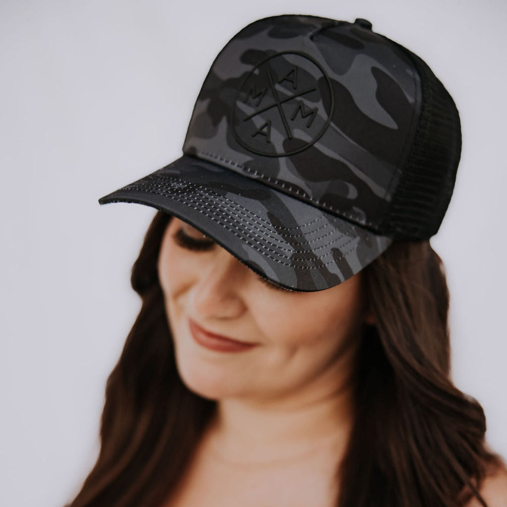 A woman with long brown hair smiles while wearing the Black Camo Mama X™ Trucker hat by Mama X™ Brand. Featuring a rubber logo design on the front, she pairs it with a black top against a plain white background. The hat includes an adjustable snapback closure for comfort.