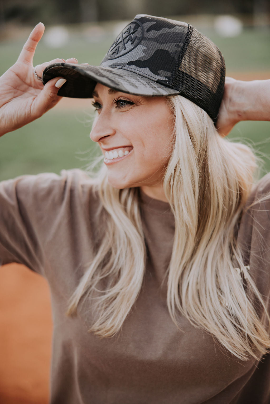 A smiling woman wears a Black Camo Mama X™ Trucker cap with an adjustable snapback and a brown T-shirt. She stands on a baseball field, slightly adjusting her cap. The blurred background shows green grass and dirt.