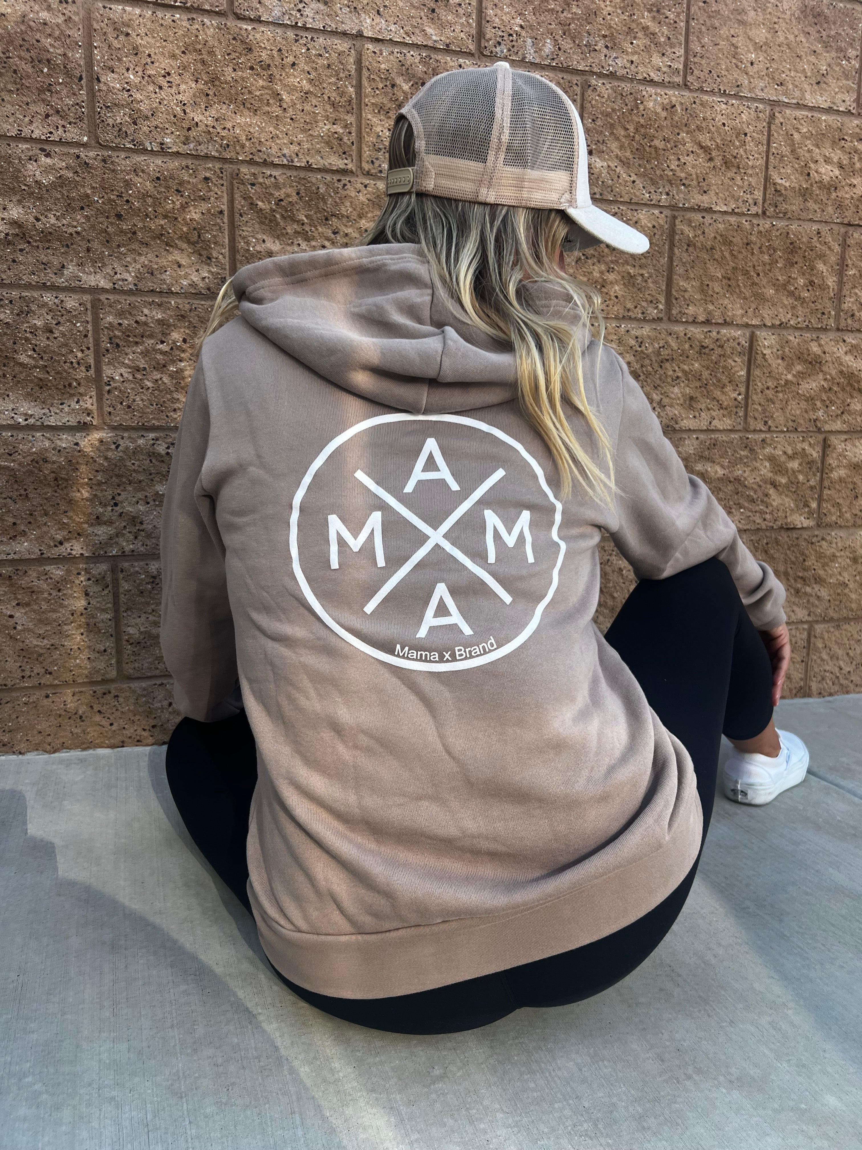 A person wearing a Mama X™ Zip Up Sweatshirt in brown, featuring "MAMA" and the "Mama X™ Brand" text on the back, is sitting against a brick wall. They have long hair and are also wearing a beige baseball cap, black leggings, and white sneakers. The hoodie is crafted from premium cotton for extra comfort.