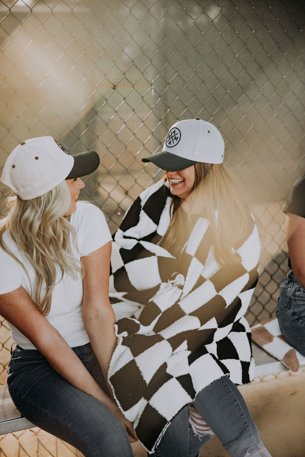 Two women in baseball caps sit smiling on a bench outdoors by a chain-link fence, one wrapped in the cozy Mama X Checkered Blanket by Mama X™ Brand—a perfect gift for Mama that brightens any moment with warmth and cheer.