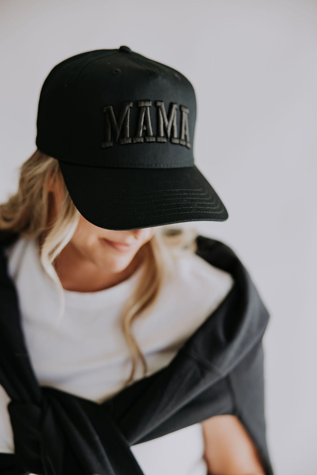 A person with long blonde hair wears the Mama X™ Brand Mama black on black Trucker Hat with 3D embroidery. Their face is partly hidden by the cap, paired with a white shirt and black jacket over their shoulders against a plain background.