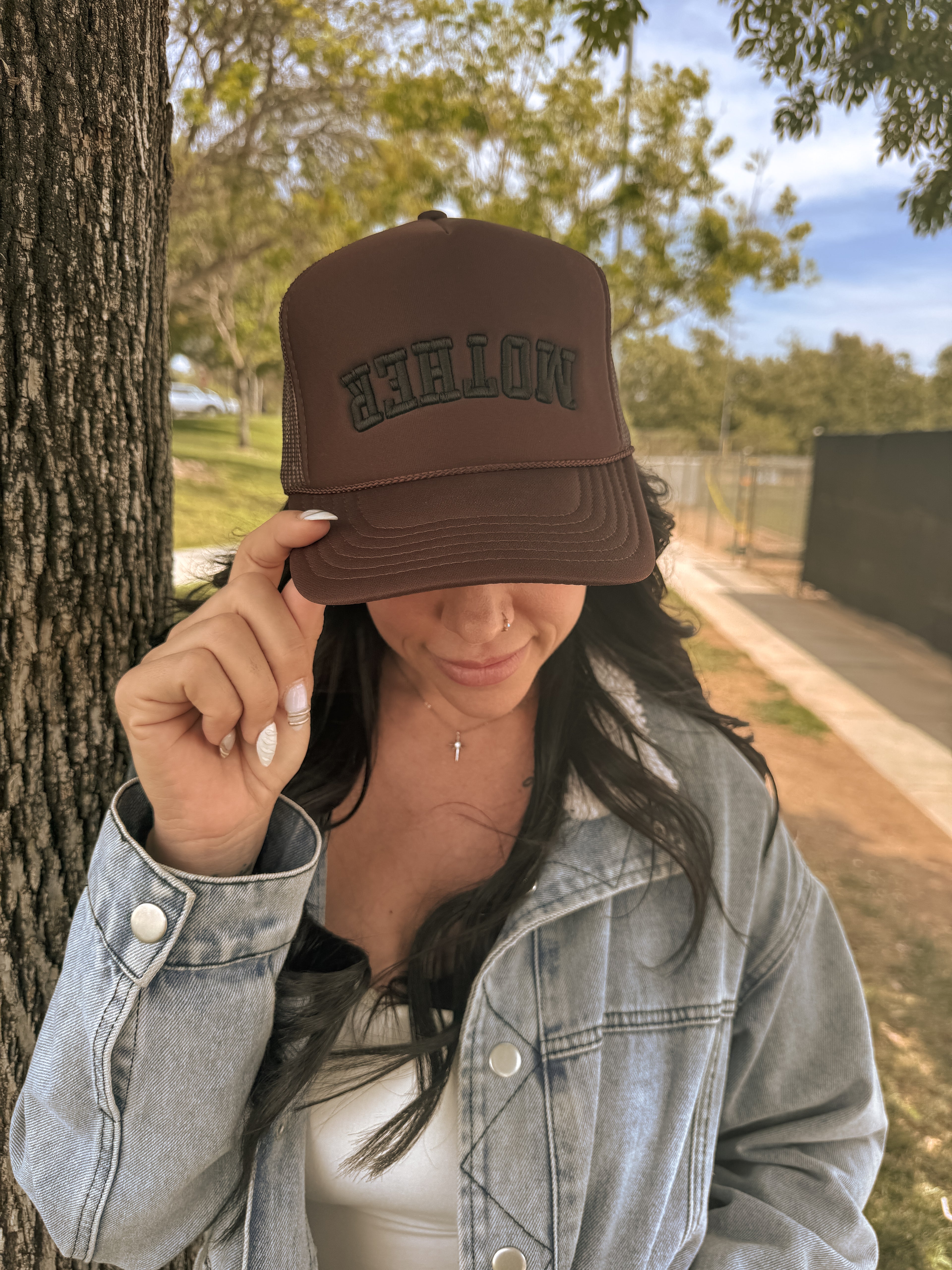 A woman outdoors tips a brown Mother Foam Trucker hat by Mama X™ Brand. With long dark hair, a light blue denim jacket, and white top, she stands beside a tree on a sunny day.