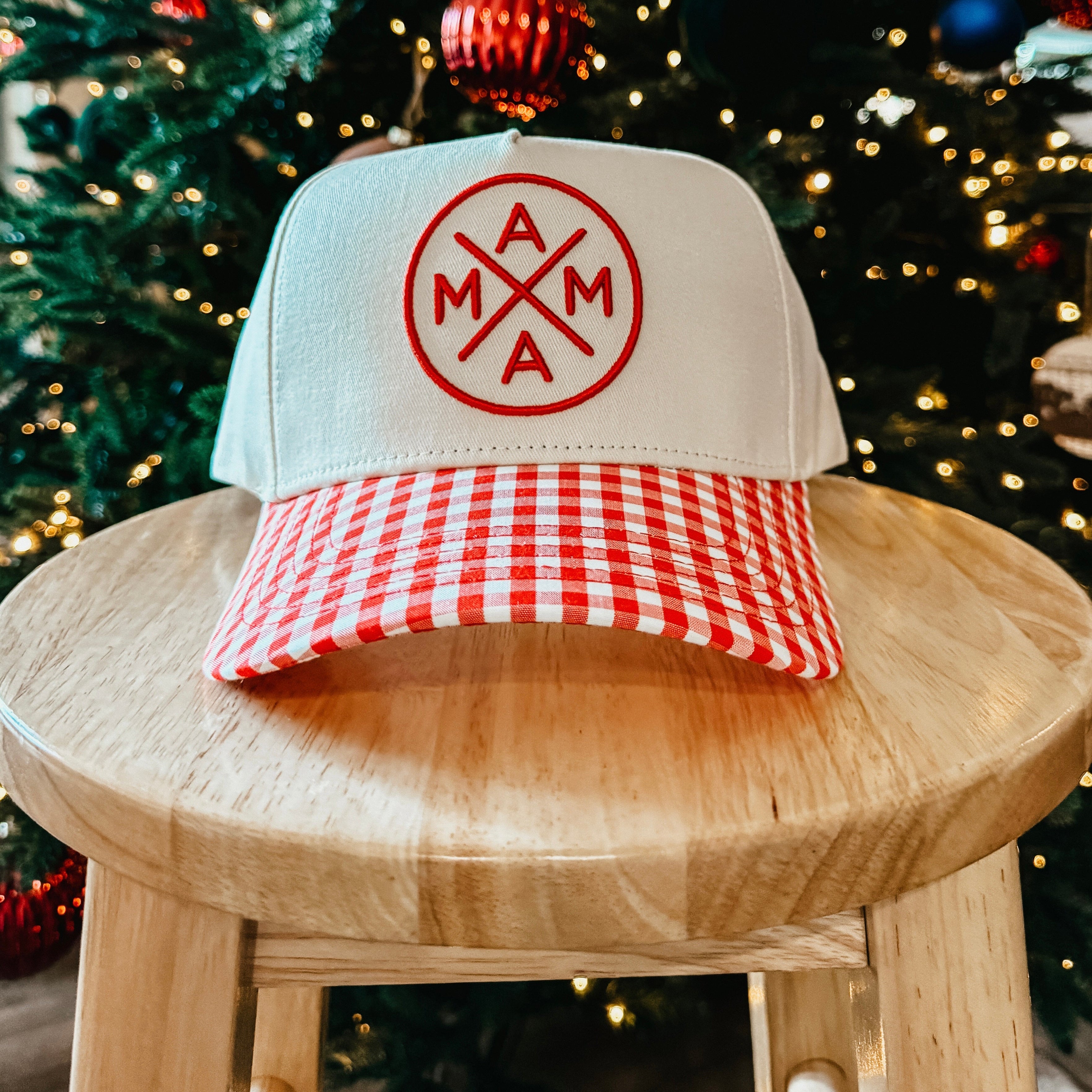 Trucker hat with a red and white checkered brim and logo on a wooden stool in front of a decorated Christmas tree.