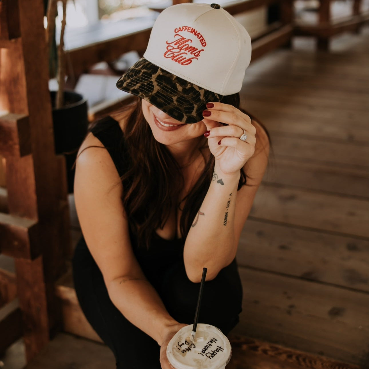 A woman in black sits on a wooden floor, smiling as she holds the brim of her Mama X™ Brand Caffeine Club Trucker cap. She sports red nail polish and sips from a cold drink with a straw.